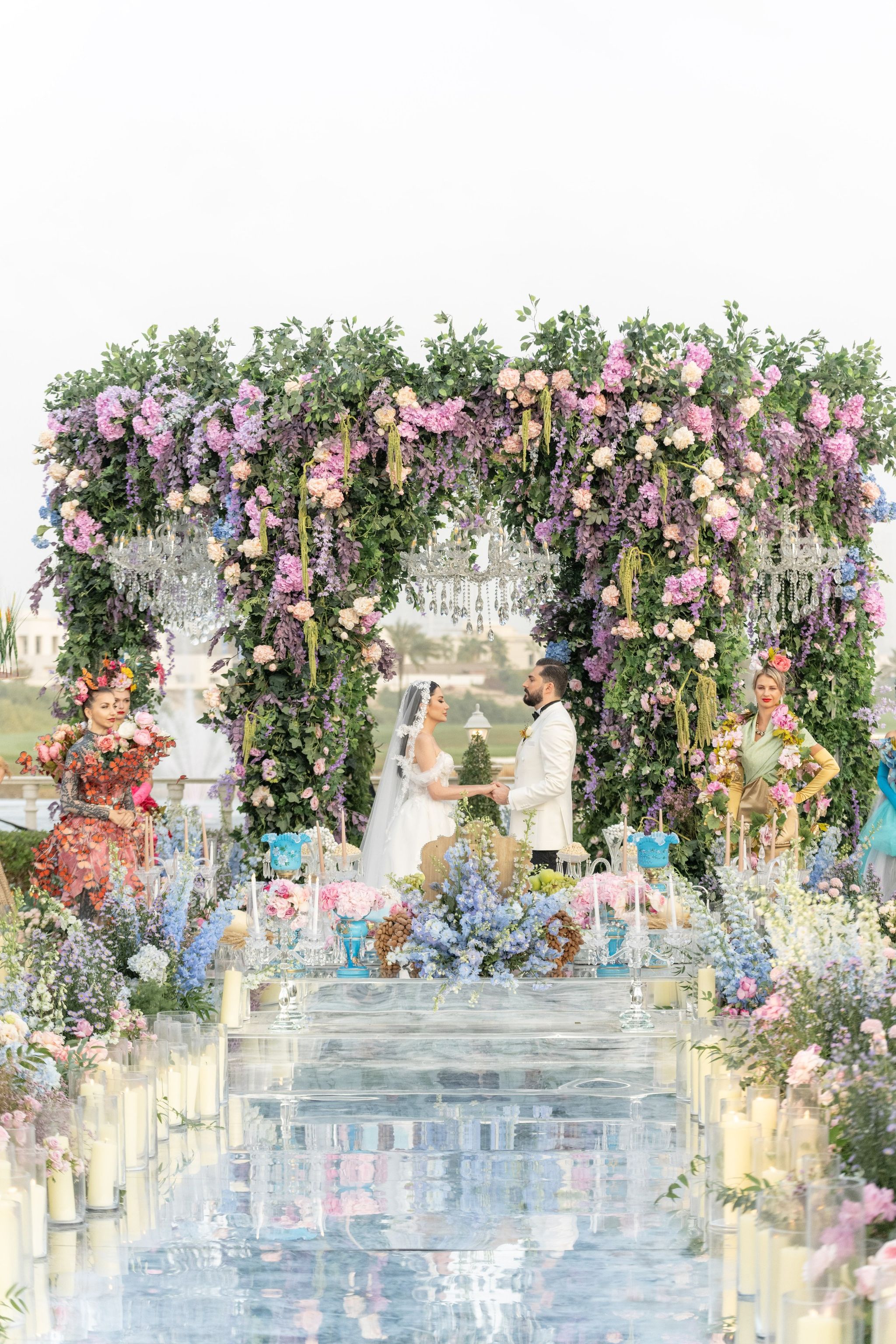 The couple holding hands beneath a chandelier-hung canopy at the end of the mirrored aisle