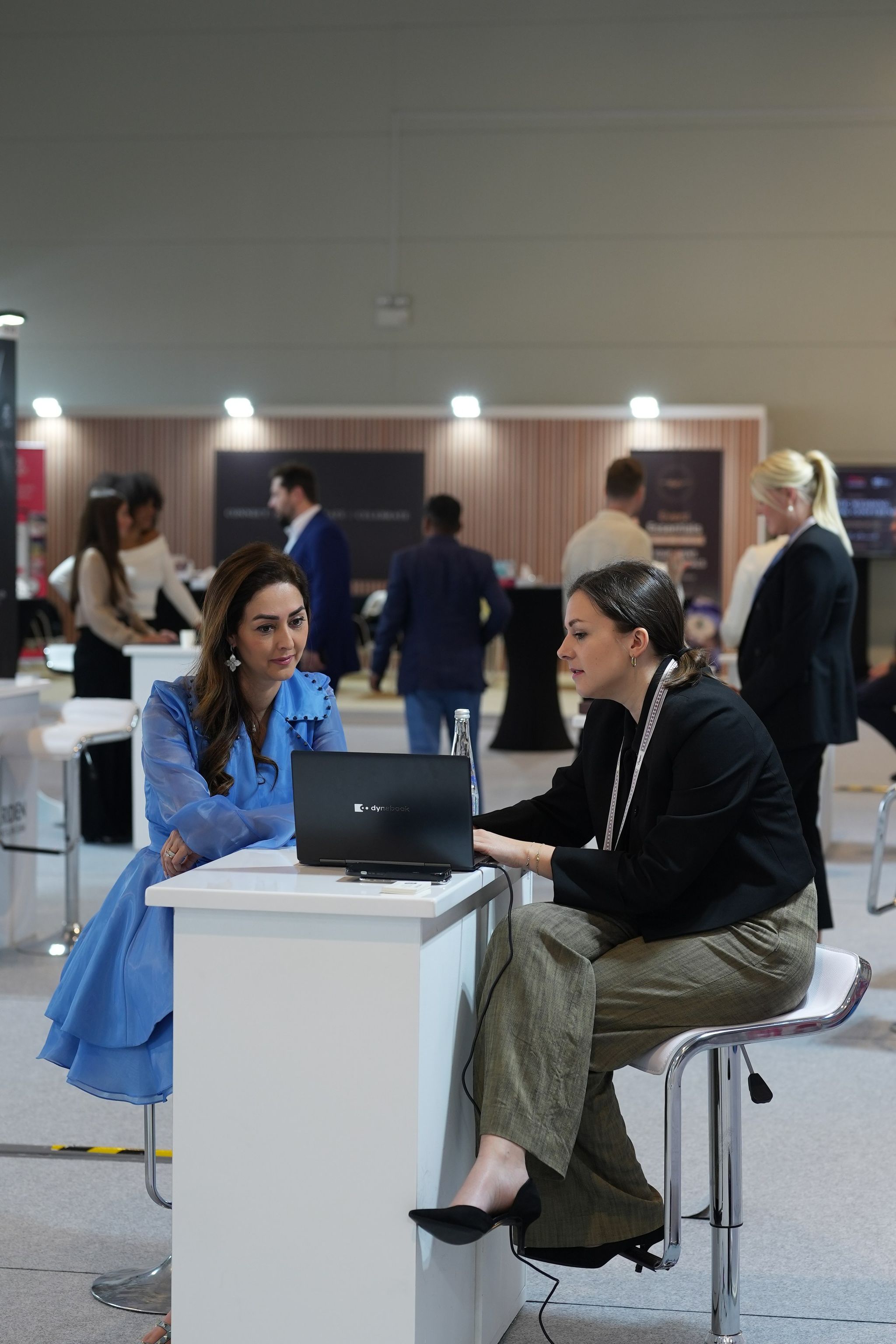 Two women meeting at a white high table with a laptop, exhibition stands behind
