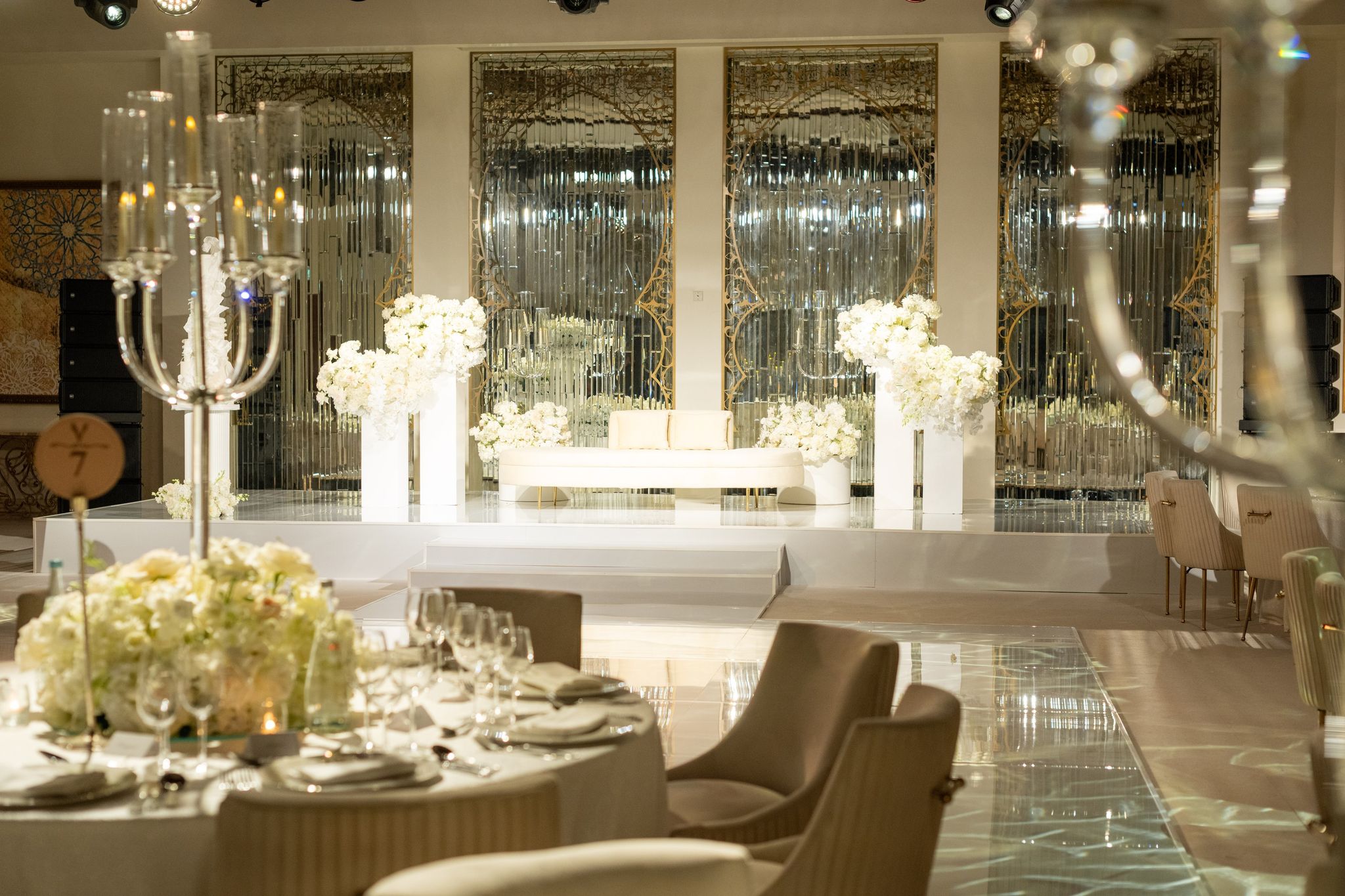 Evening reception stage with a white loveseat and floral plinths before mirrored, gold-framed panels