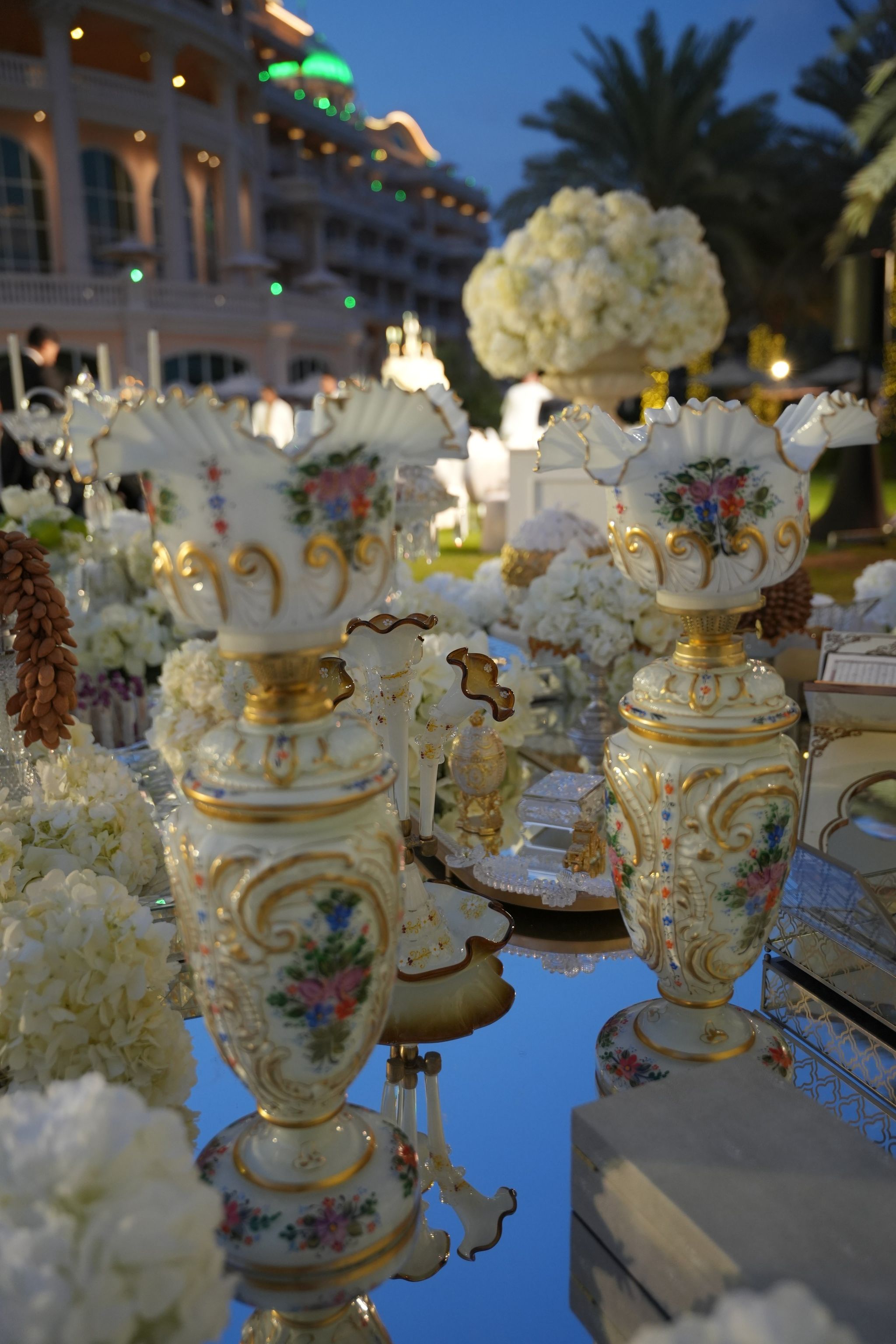 Hand-painted porcelain vases on a mirrored sofreh at dusk, with the illuminated facade of Raffles The Palm Dubai behind