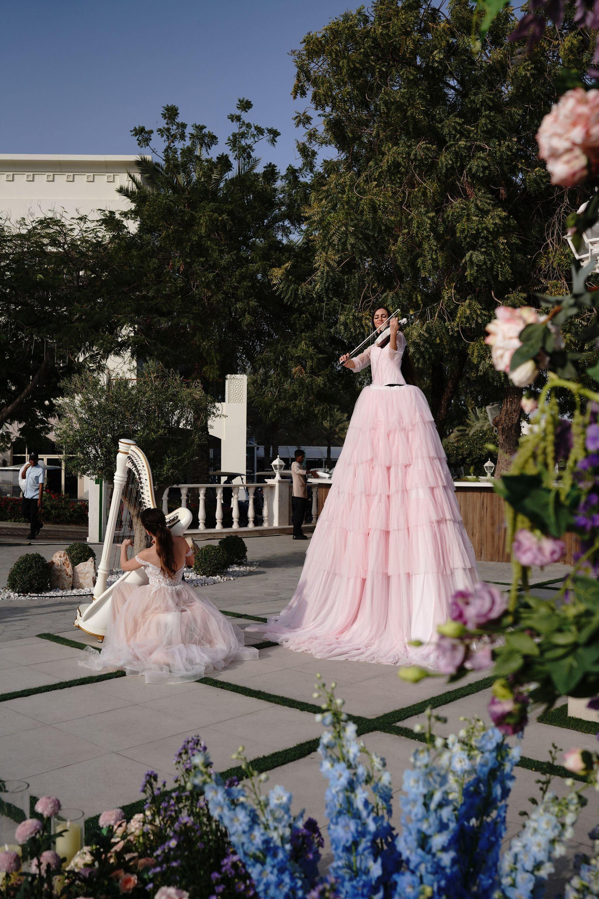 Stilt violinist and harpist in blush tulle gowns, framed by blue delphinium and roses