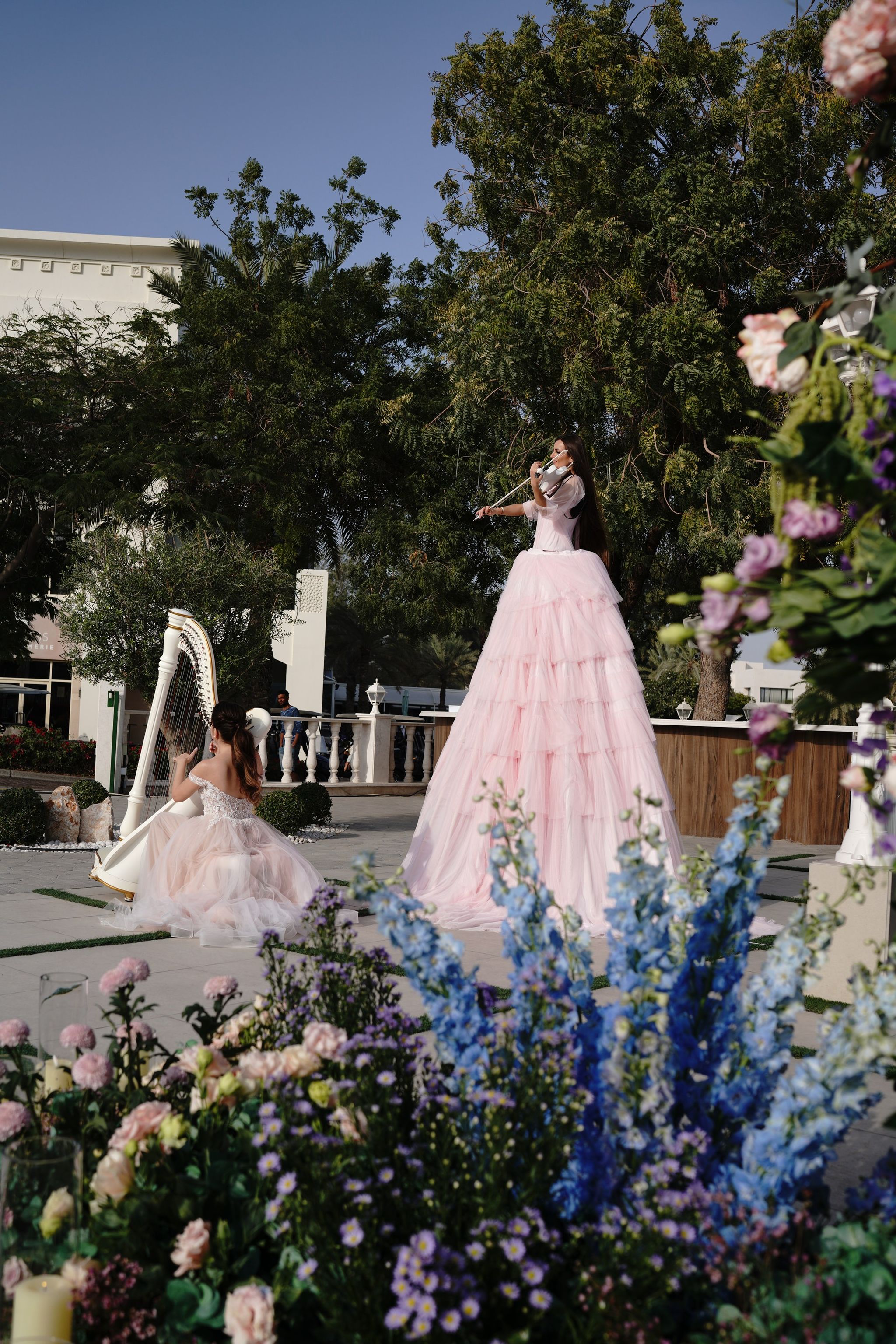 Violinist in a towering pink gown and a harpist, seen beyond delphinium and garden roses