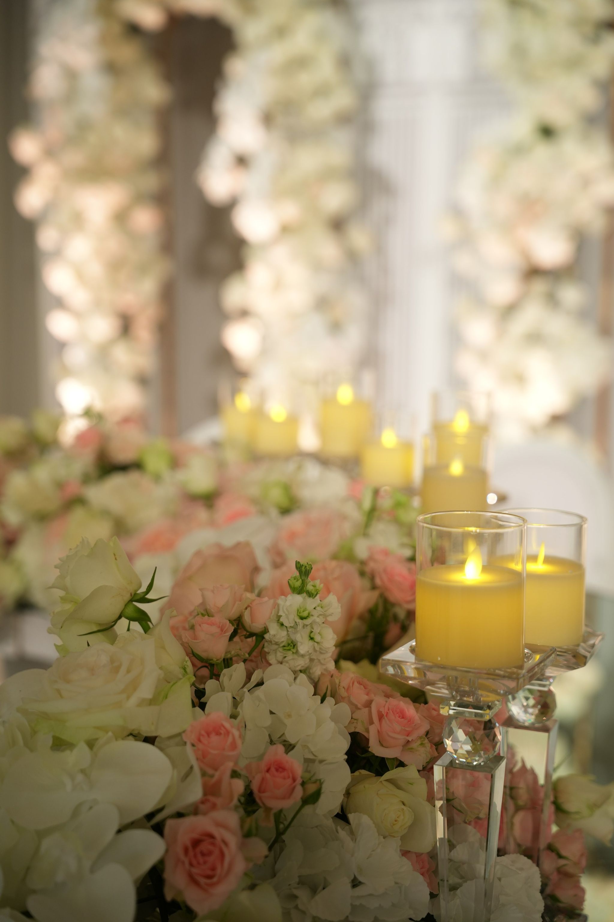 Pillar candles in crystal holders among peach roses and white orchids, arches glowing behind