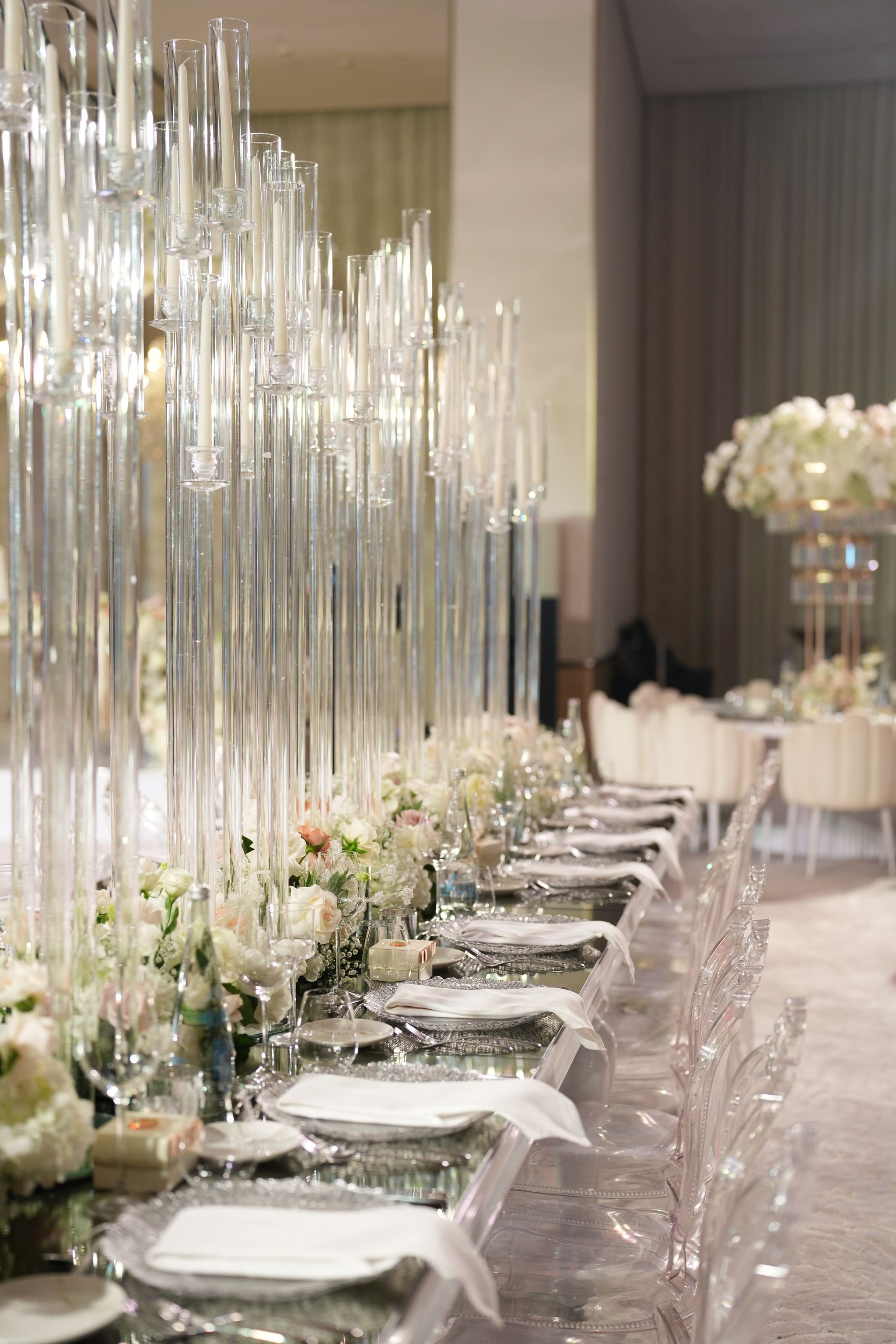 Rows of tall glass candle columns over blush roses and folded napkins on a mirrored table