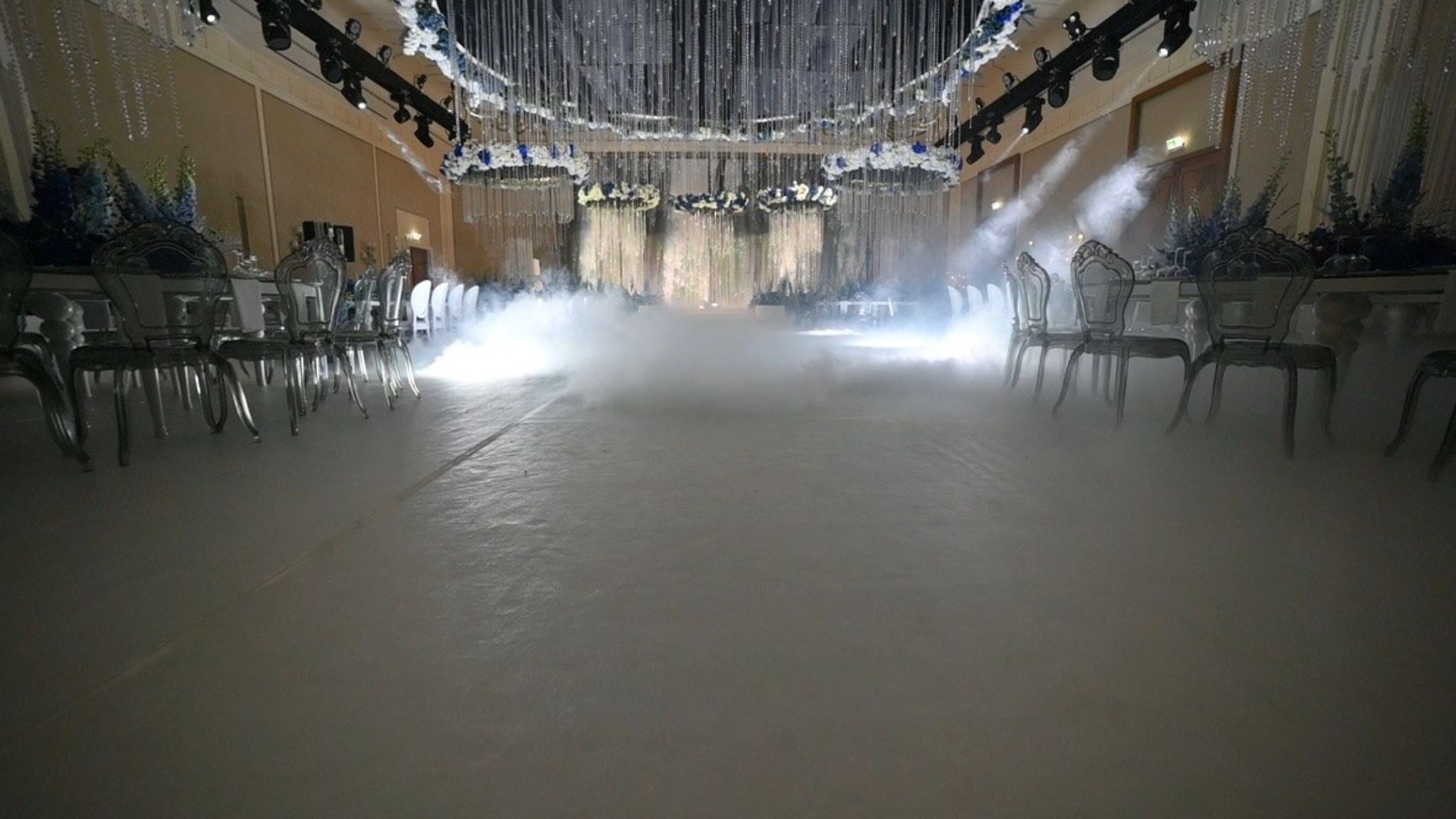 Low fog rolling across the ballroom floor between clear chairs towards the floral rings