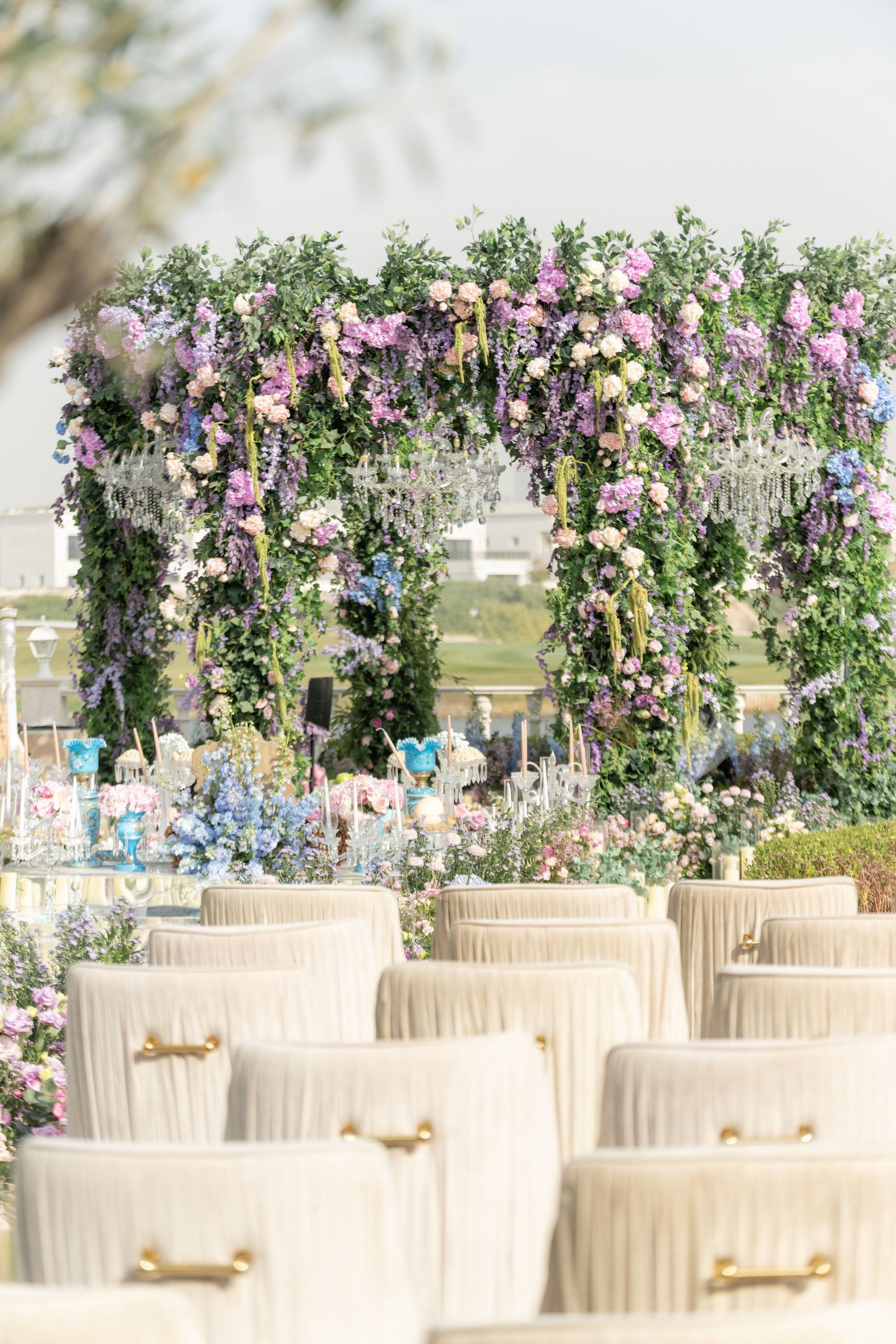 Rows of pleated ivory chairs with brass handles before a lilac and blush floral canopy