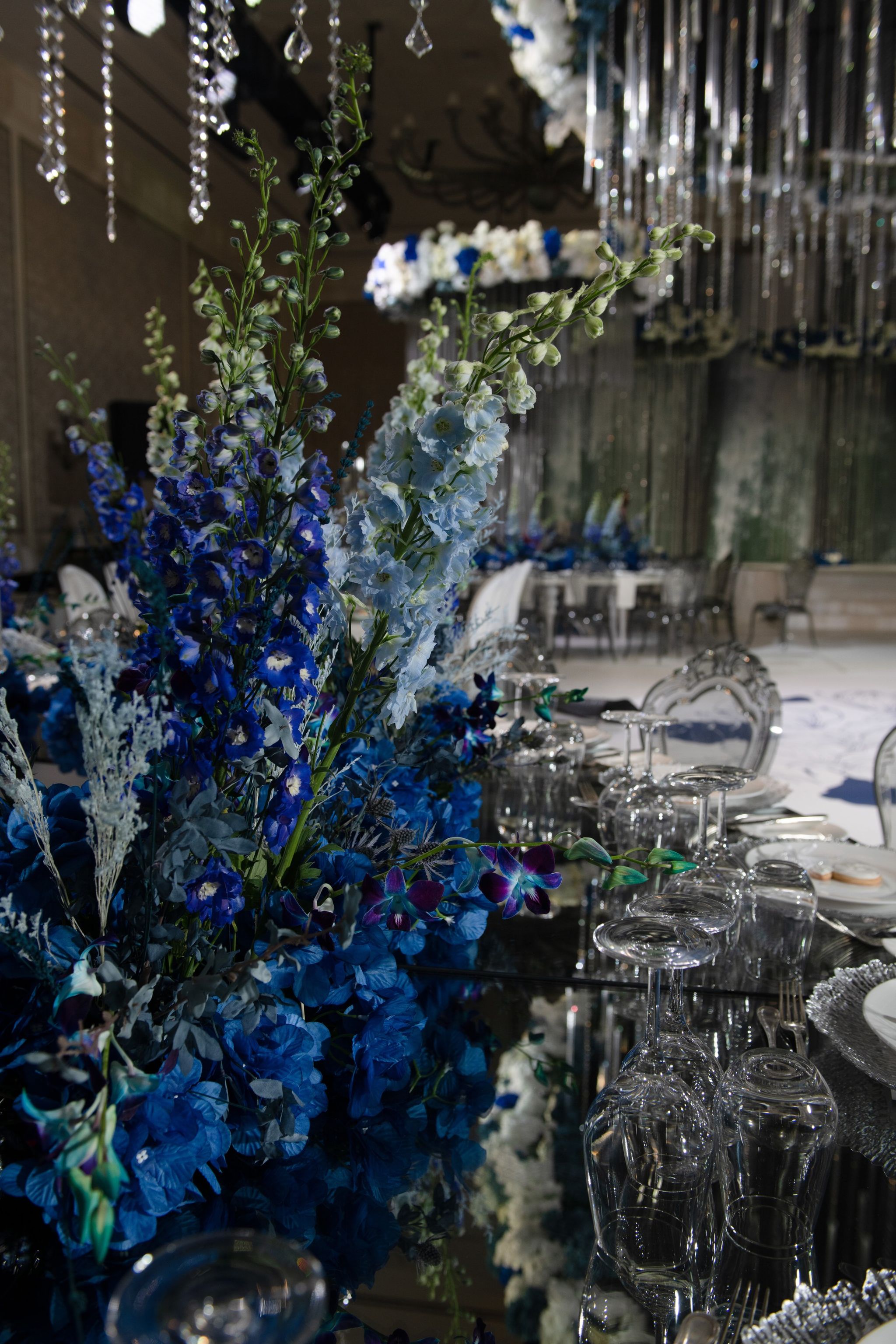 Blue delphiniums, hydrangeas and orchids rising from a mirrored table set with crystal glassware