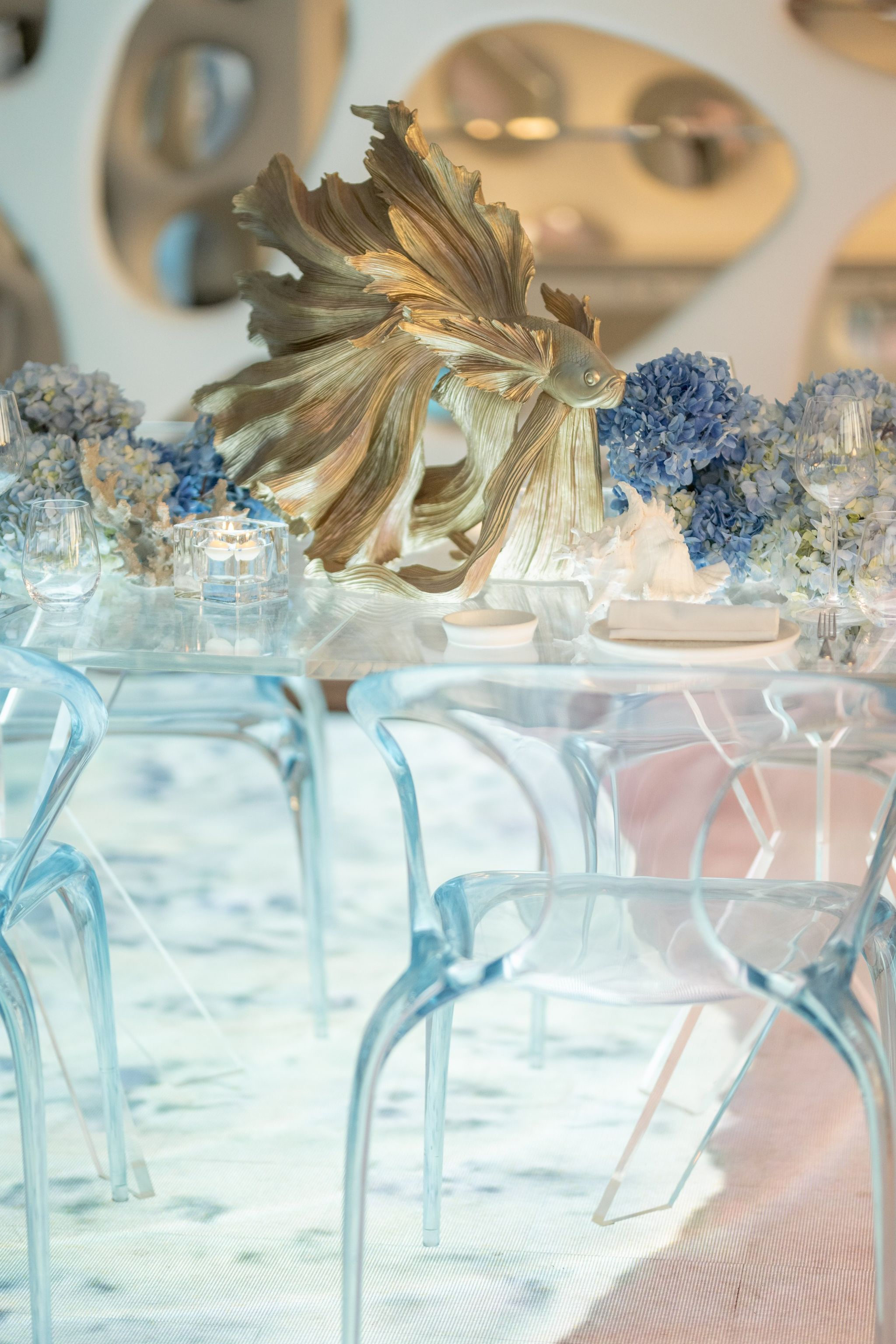 Gilded betta fish sculpture among blue hydrangeas on a clear table with acrylic chairs