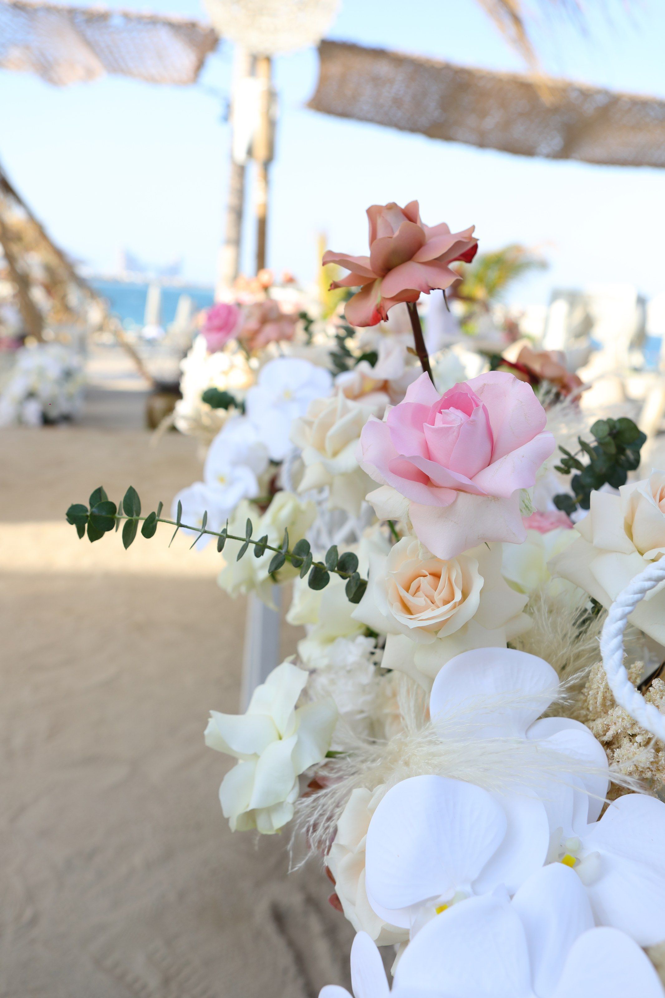 White orchids and blush roses in the foreground, a sandy path and the sea softly blurred behind