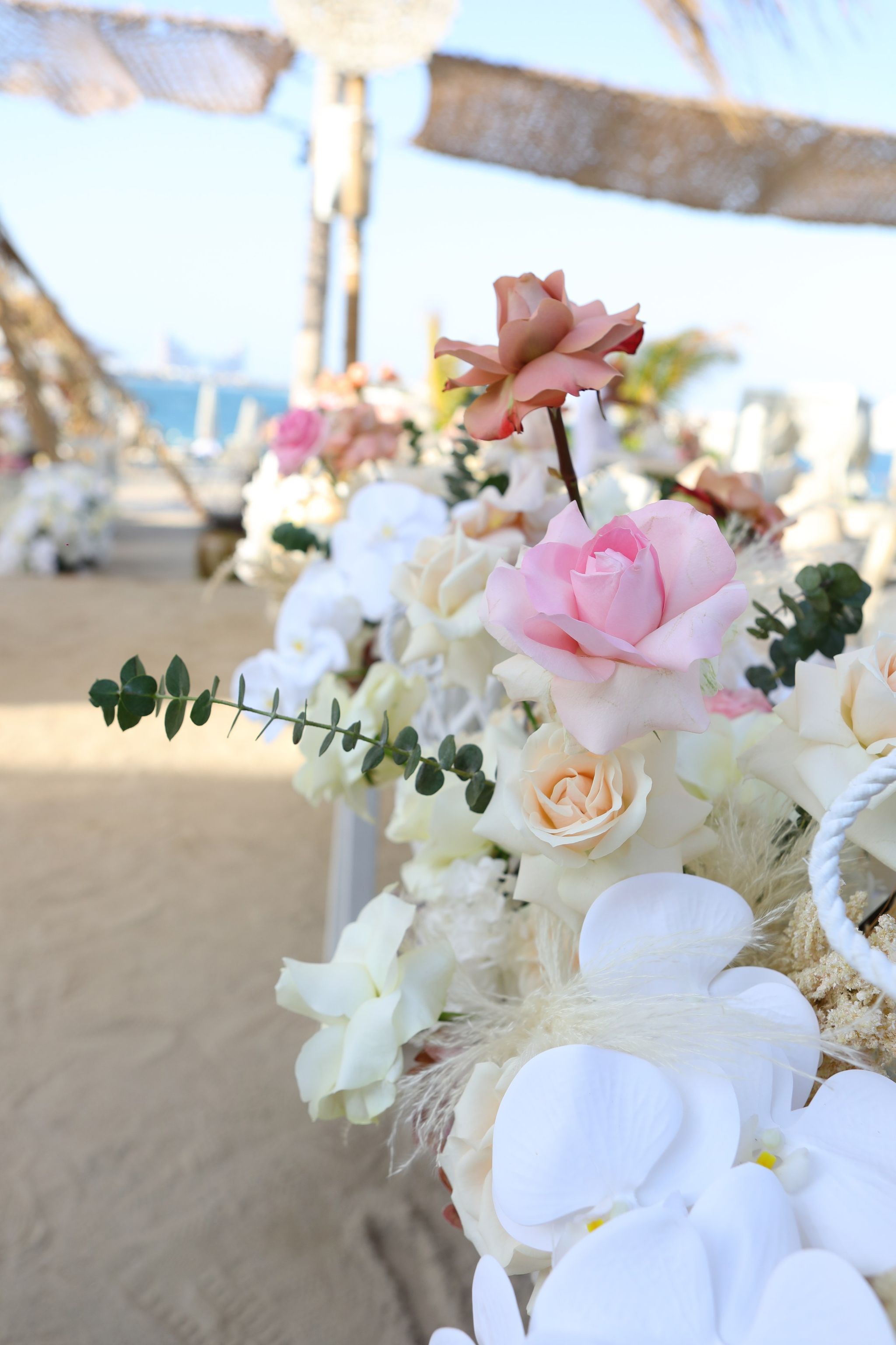 White orchids and blush roses in the foreground, a sandy path and the sea softly blurred behind