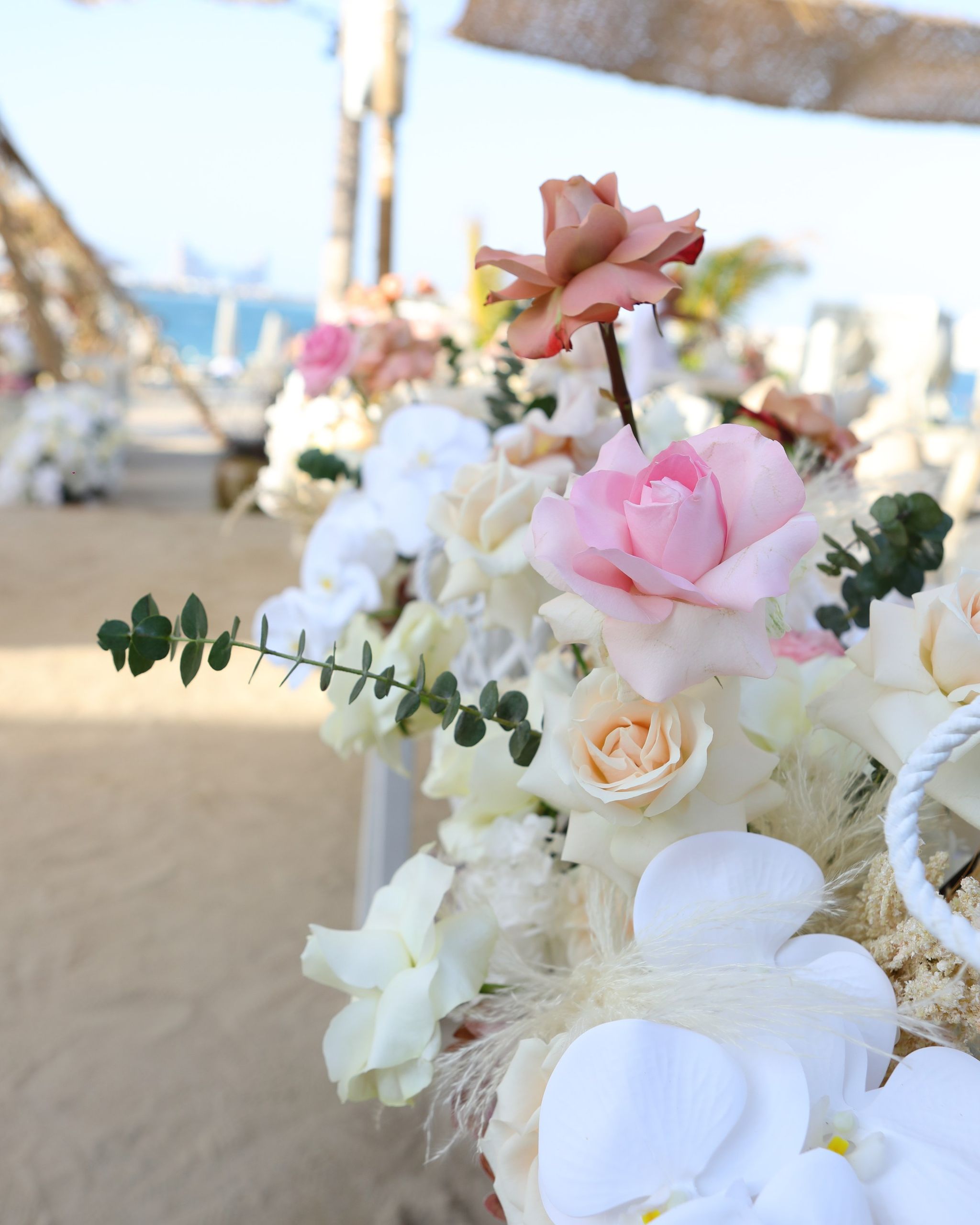 White orchids and blush roses in the foreground, a sandy path and the sea softly blurred behind