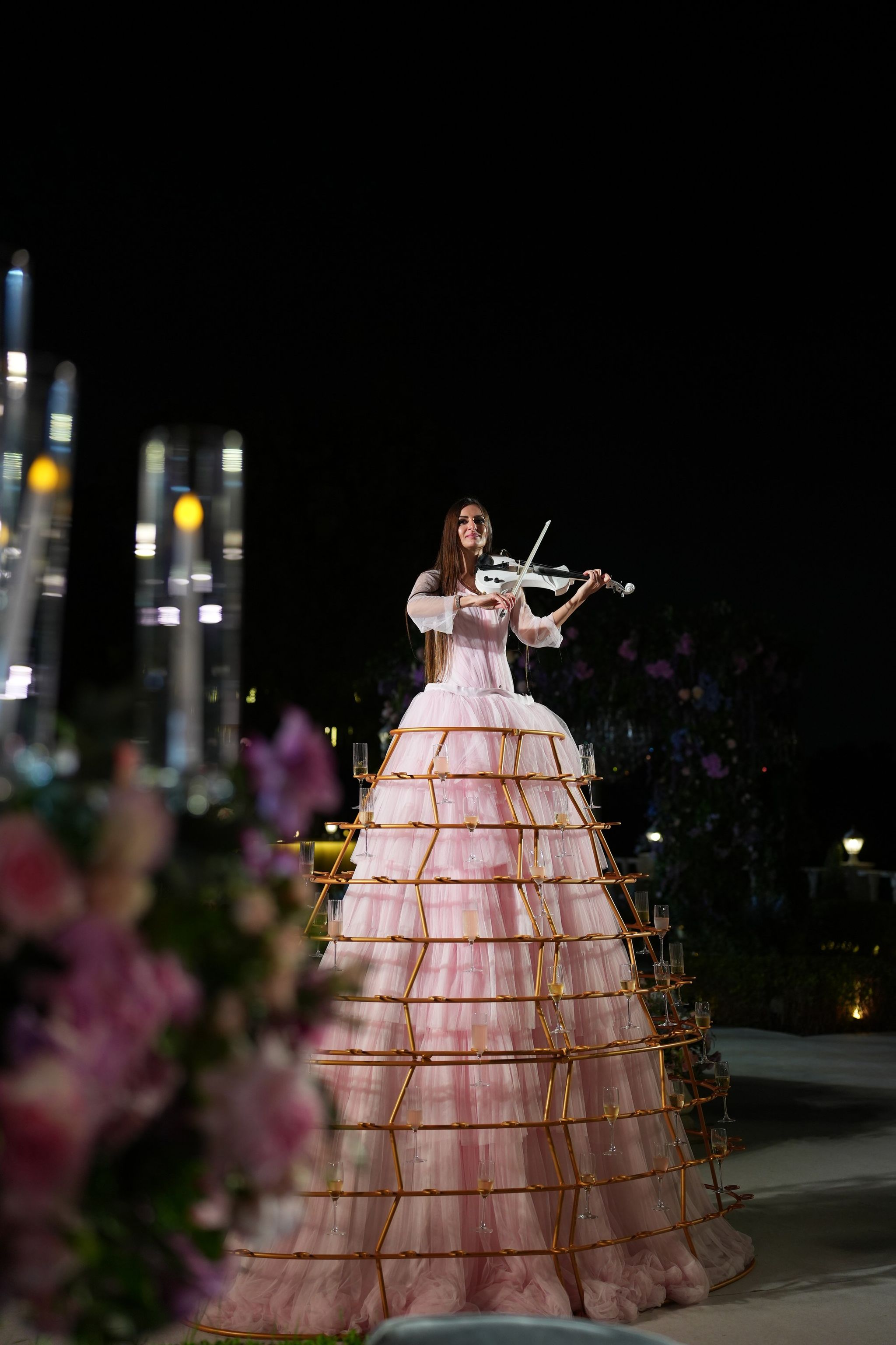 Night view of a violinist in a champagne-glass skirt beyond candles and roses