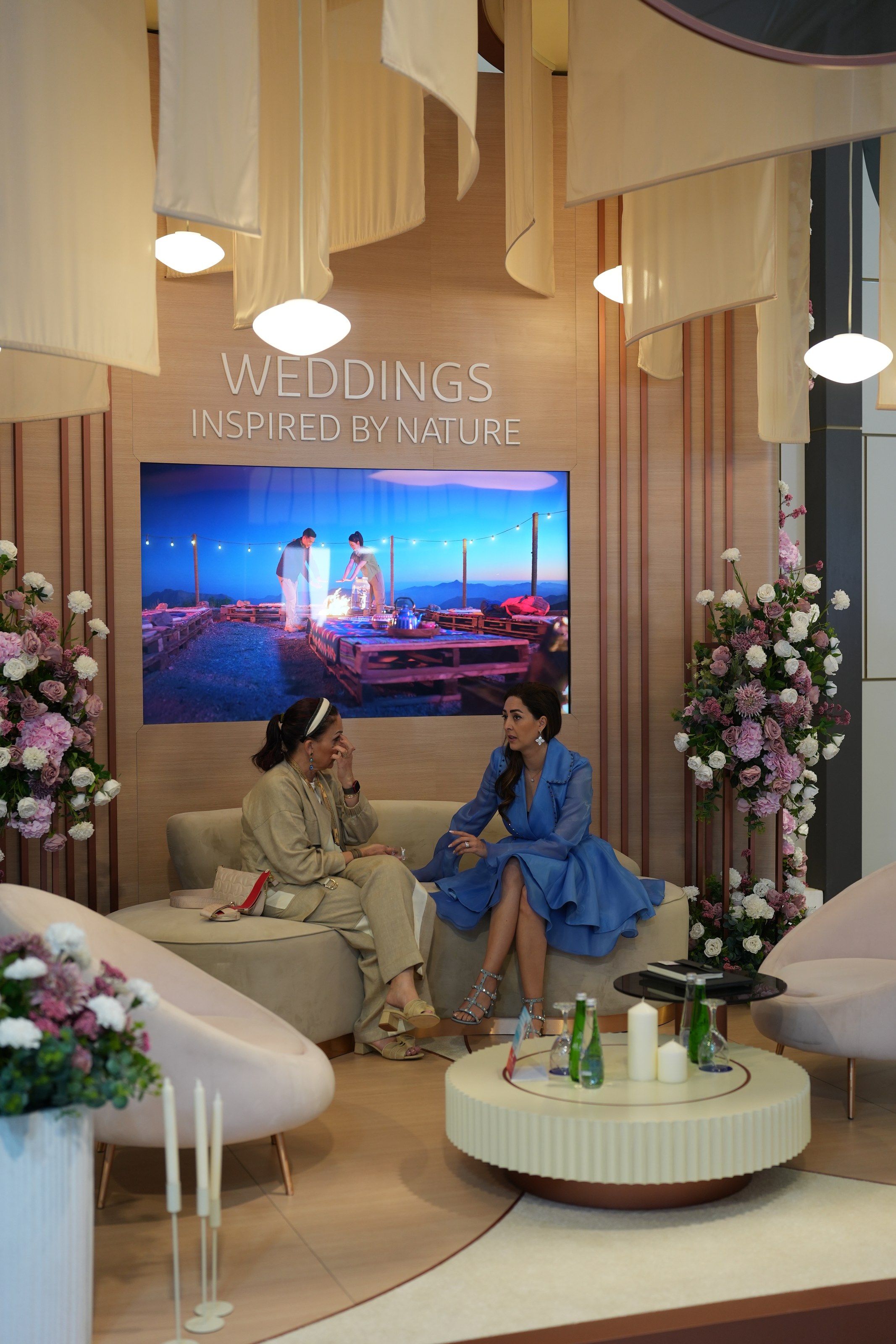 Exhibition lounge with curved blush seating, hydrangea columns and fabric canopies beneath a wedding screen