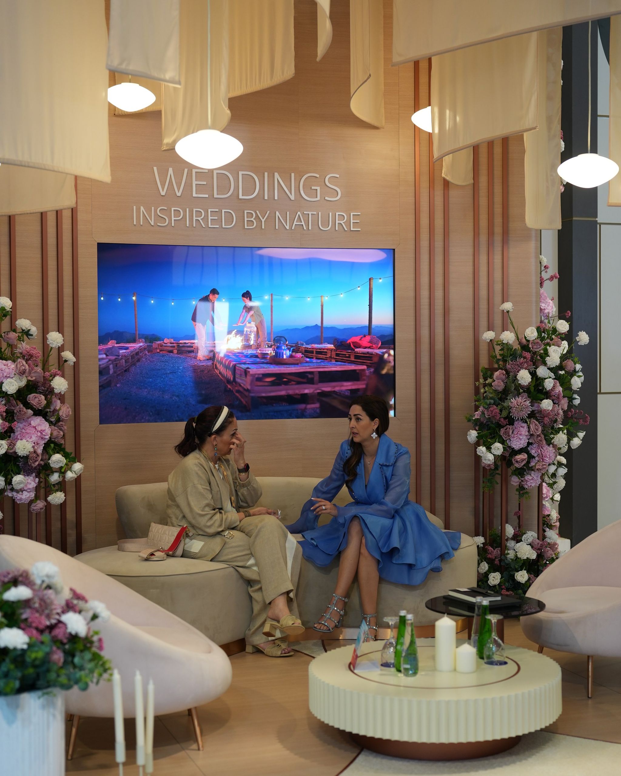Exhibition lounge with curved blush seating, hydrangea columns and fabric canopies beneath a wedding screen