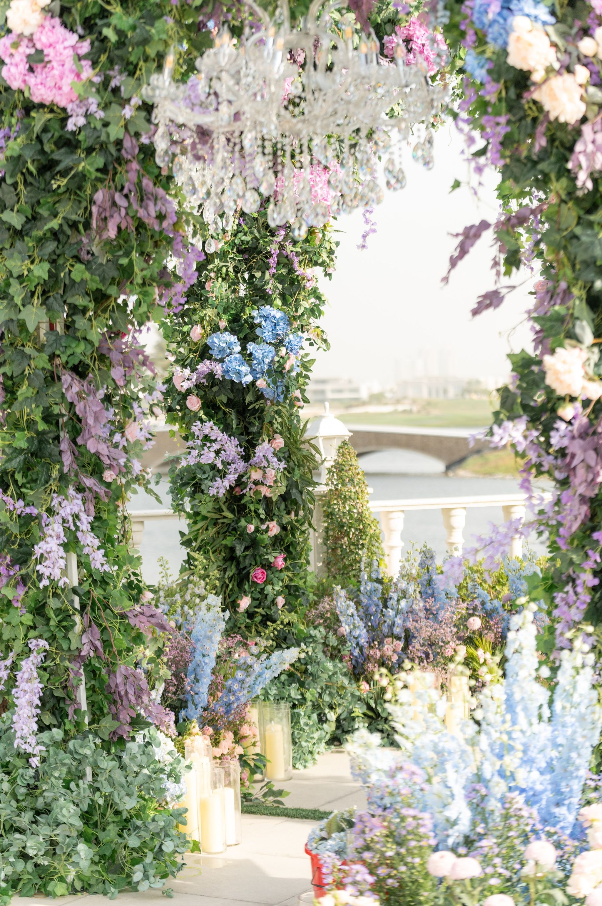 Crystal chandelier hanging within a canopy of hydrangea and wisteria, with delphinium and candles below