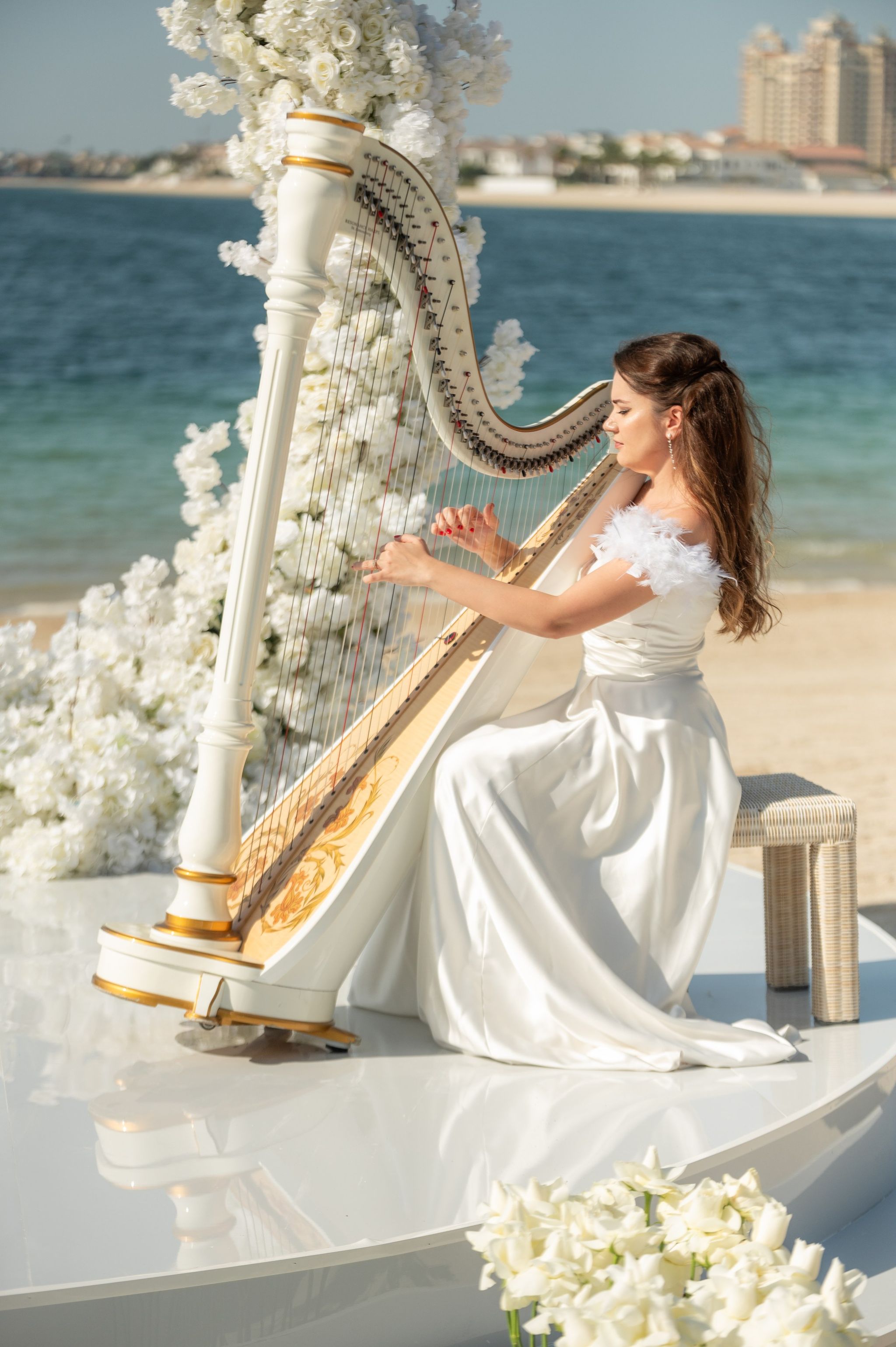Harpist in a white gown playing a gilded white harp beside the floral arch, sea behind