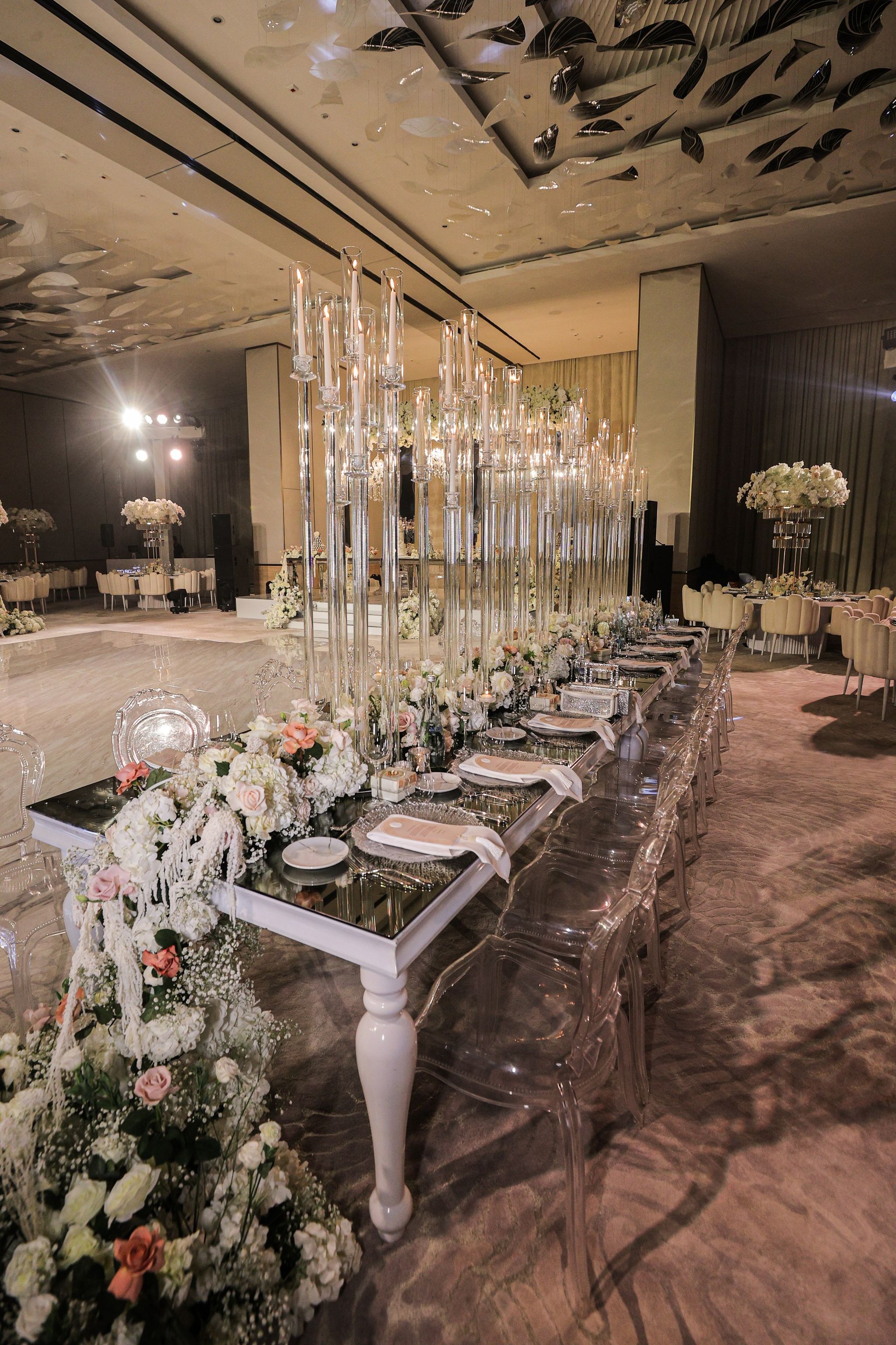 Long mirrored table with towering glass candelabra, ghost chairs and a cascading white floral arrangement