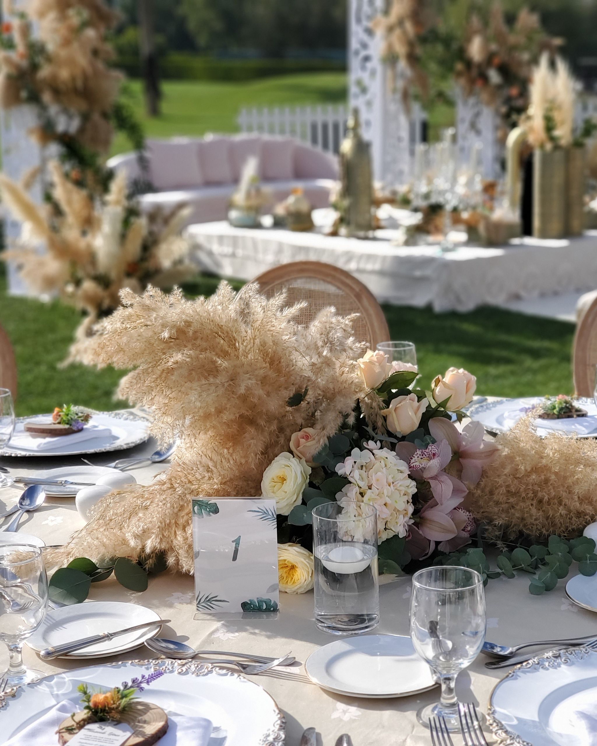 Pampas and garden rose centrepiece with table number and floating candle, ceremony lounge softly behind