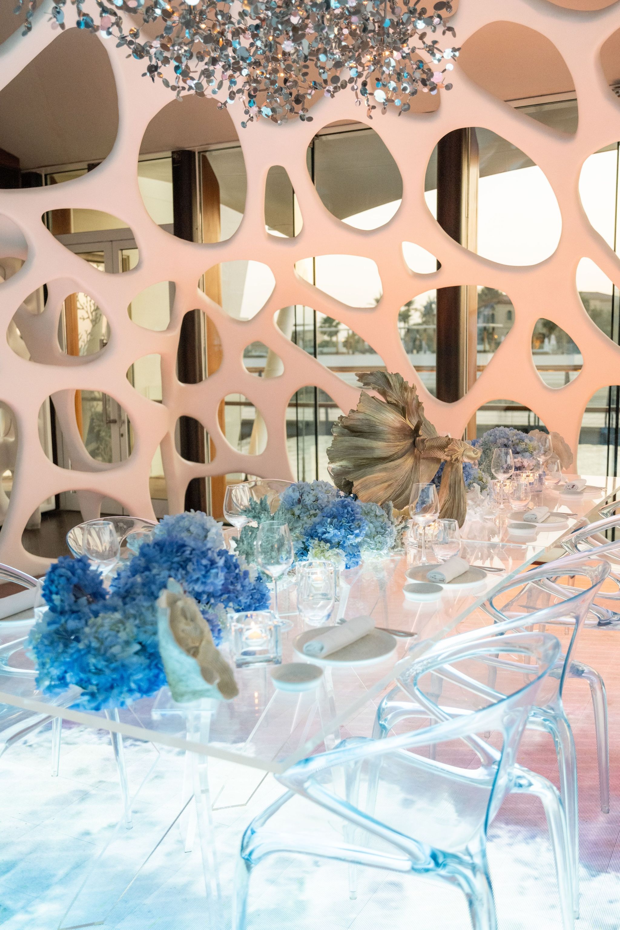 Long acrylic table of blue hydrangeas and a gilded fish sculpture beneath a crystal installation