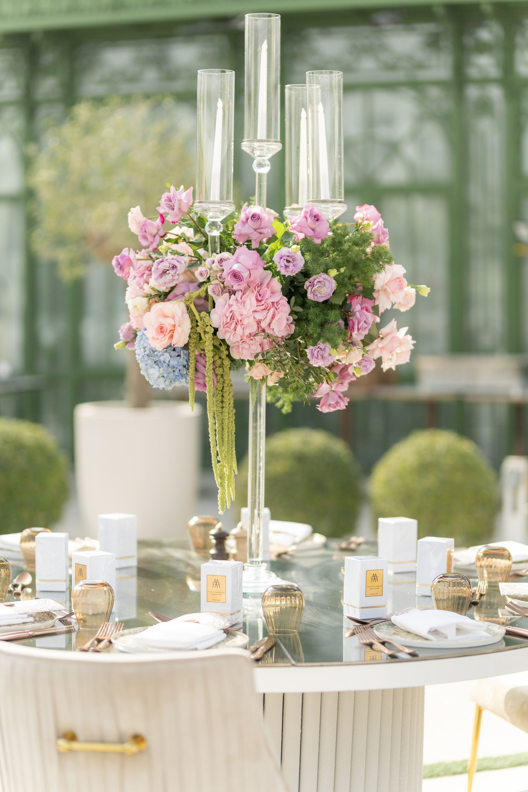 Glass candelabrum of lilac roses and hydrangea above a table set with gold-labelled favour boxes
