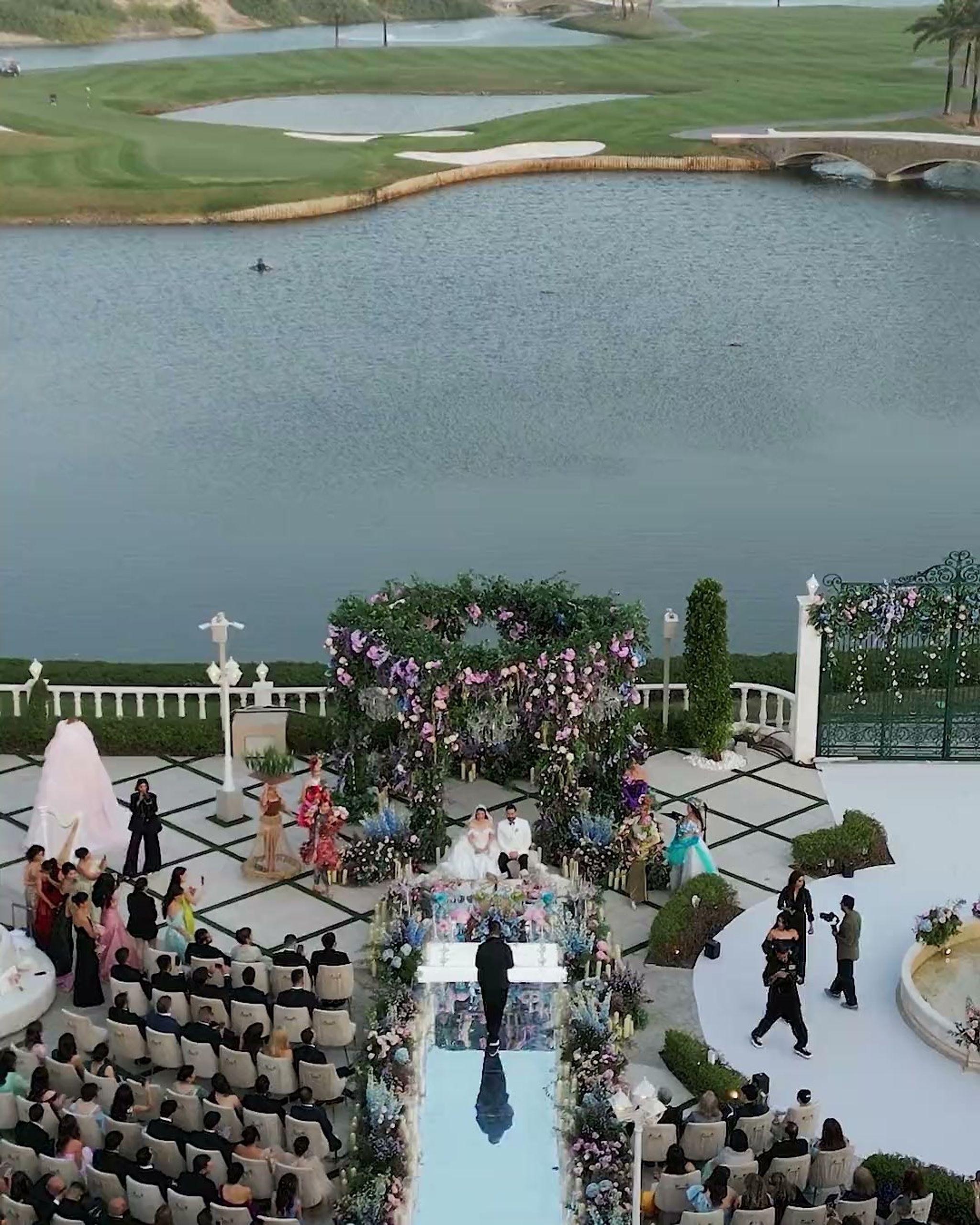 Aerial view of a lakeside terrace wedding ceremony, guests seated either side of a flower-lined mirrored aisle