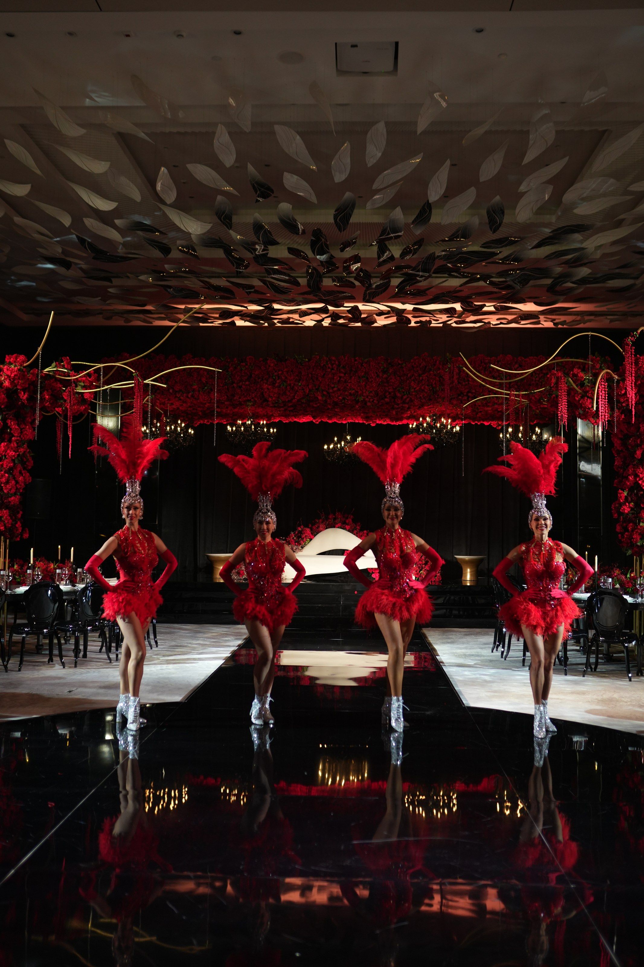 Four showgirls in red feathered headdresses on a black mirrored runway before rose-covered walls