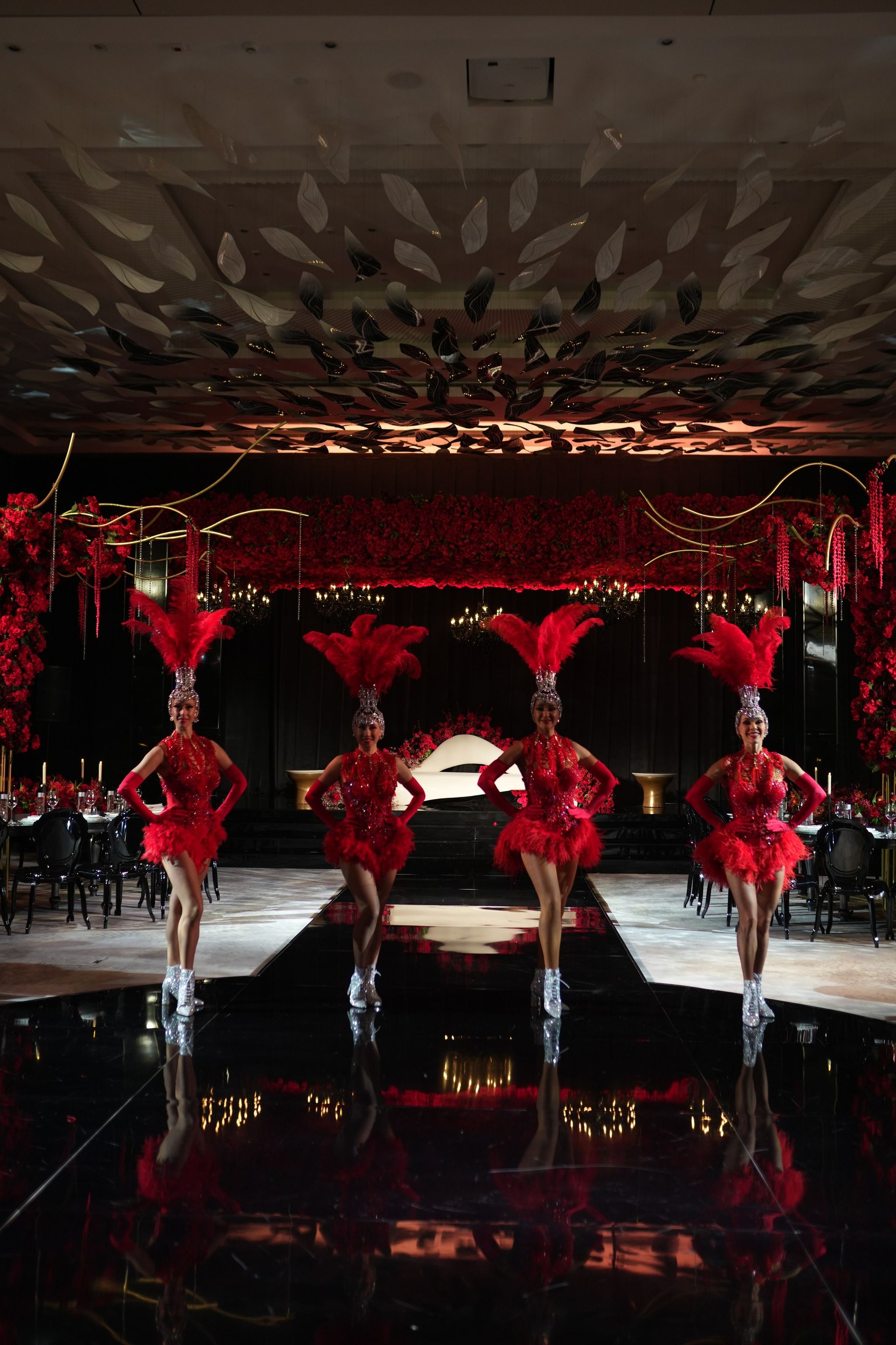Four showgirls in red feathered headdresses on a black mirrored runway before rose-covered walls