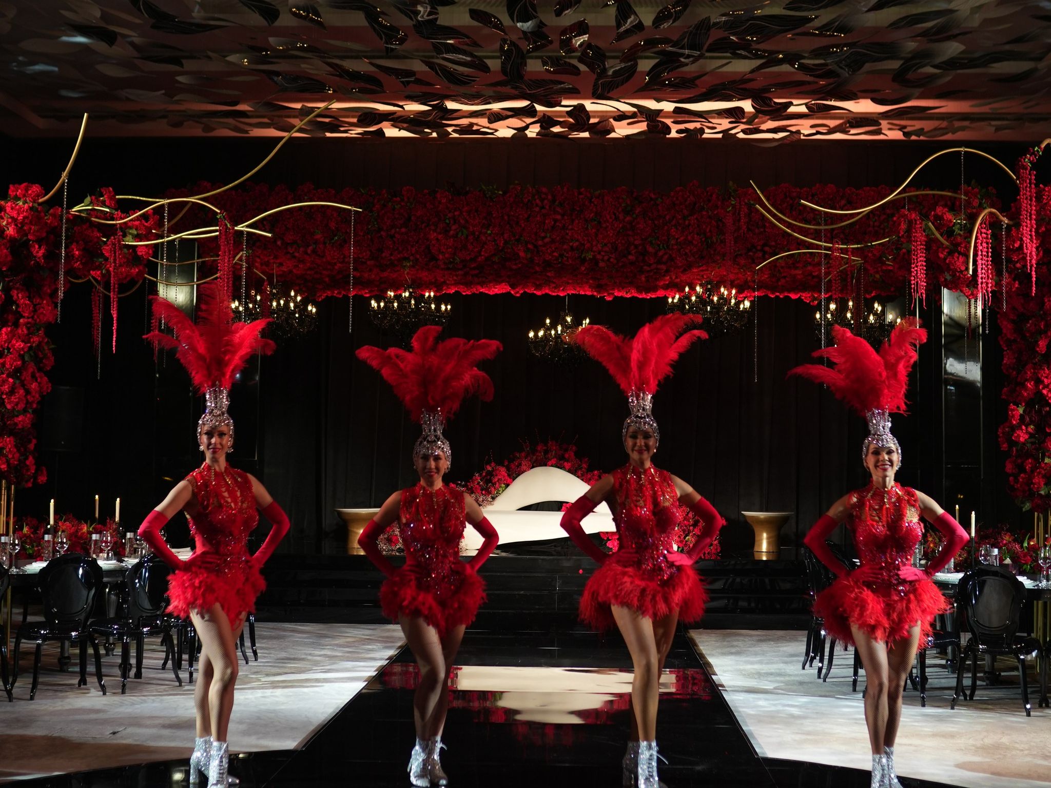 Four showgirls in red feathered headdresses on a black mirrored runway before rose-covered walls