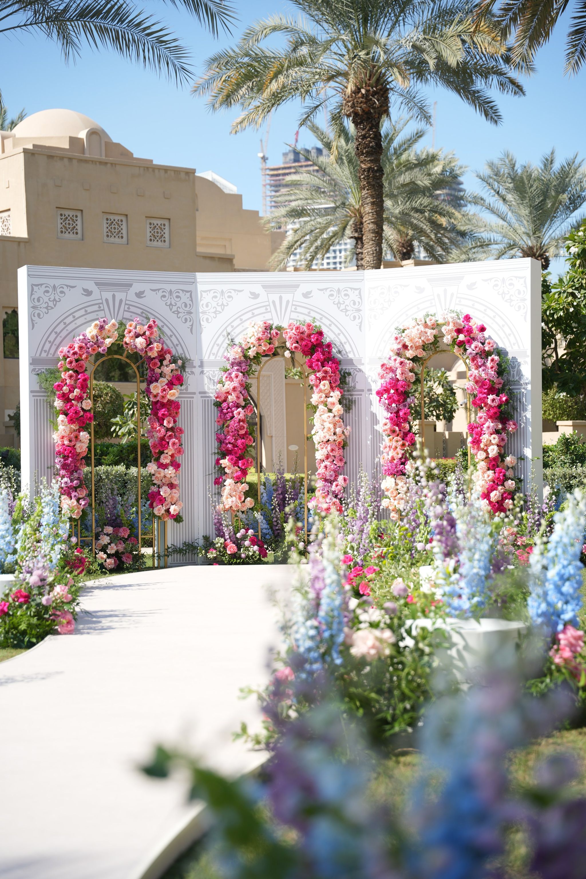 White carved ceremony screen with three rose arches beside a delphinium-lined aisle at One&Only Royal Mirage Dubai