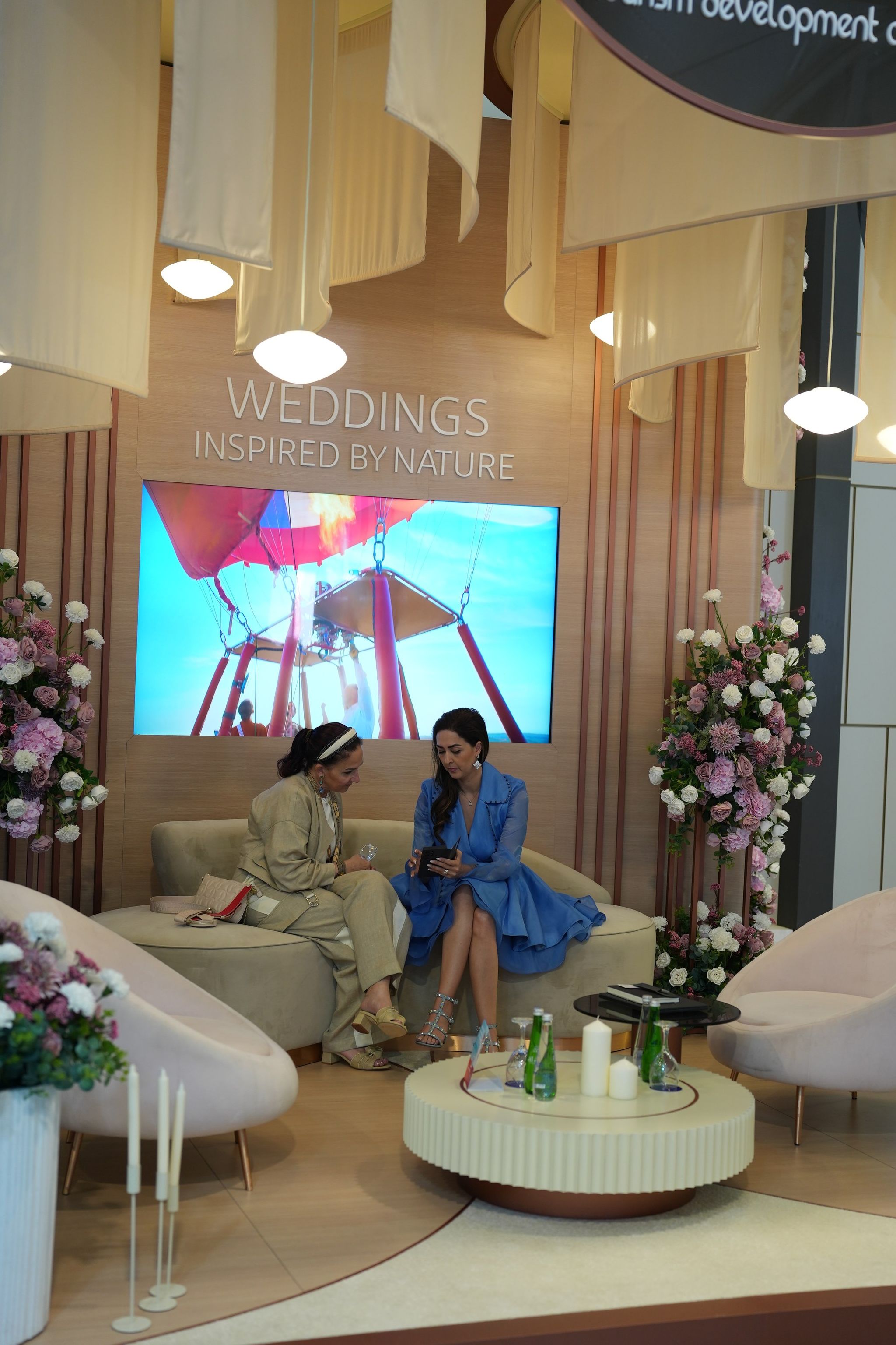 Two women in conversation on a curved sofa in a floral exhibition lounge with draped canopies