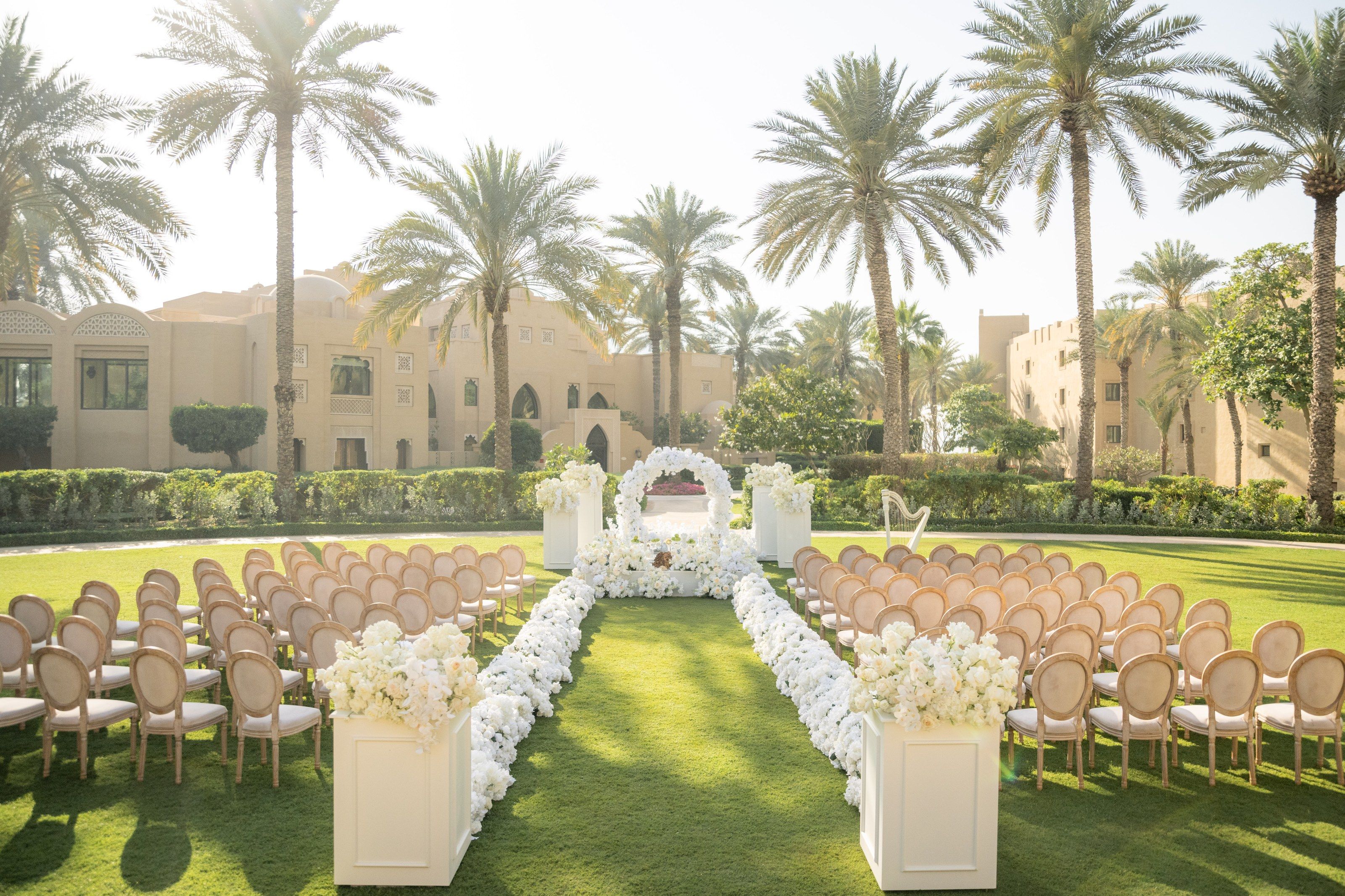 Lawn ceremony with cane-backed chairs, a white floral aisle and round arch beneath date palms
