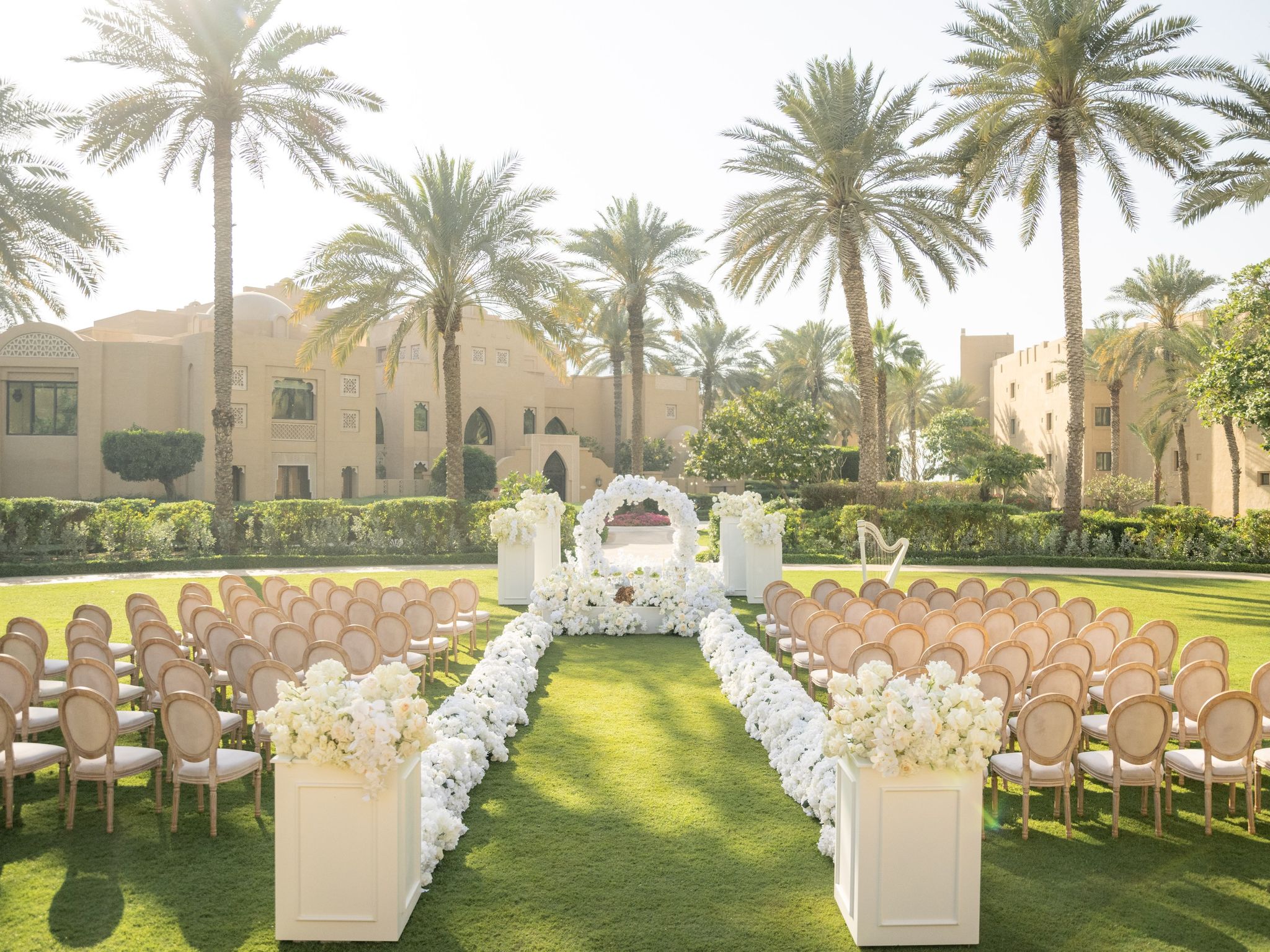 Lawn ceremony with cane-backed chairs, a white floral aisle and round arch beneath date palms
