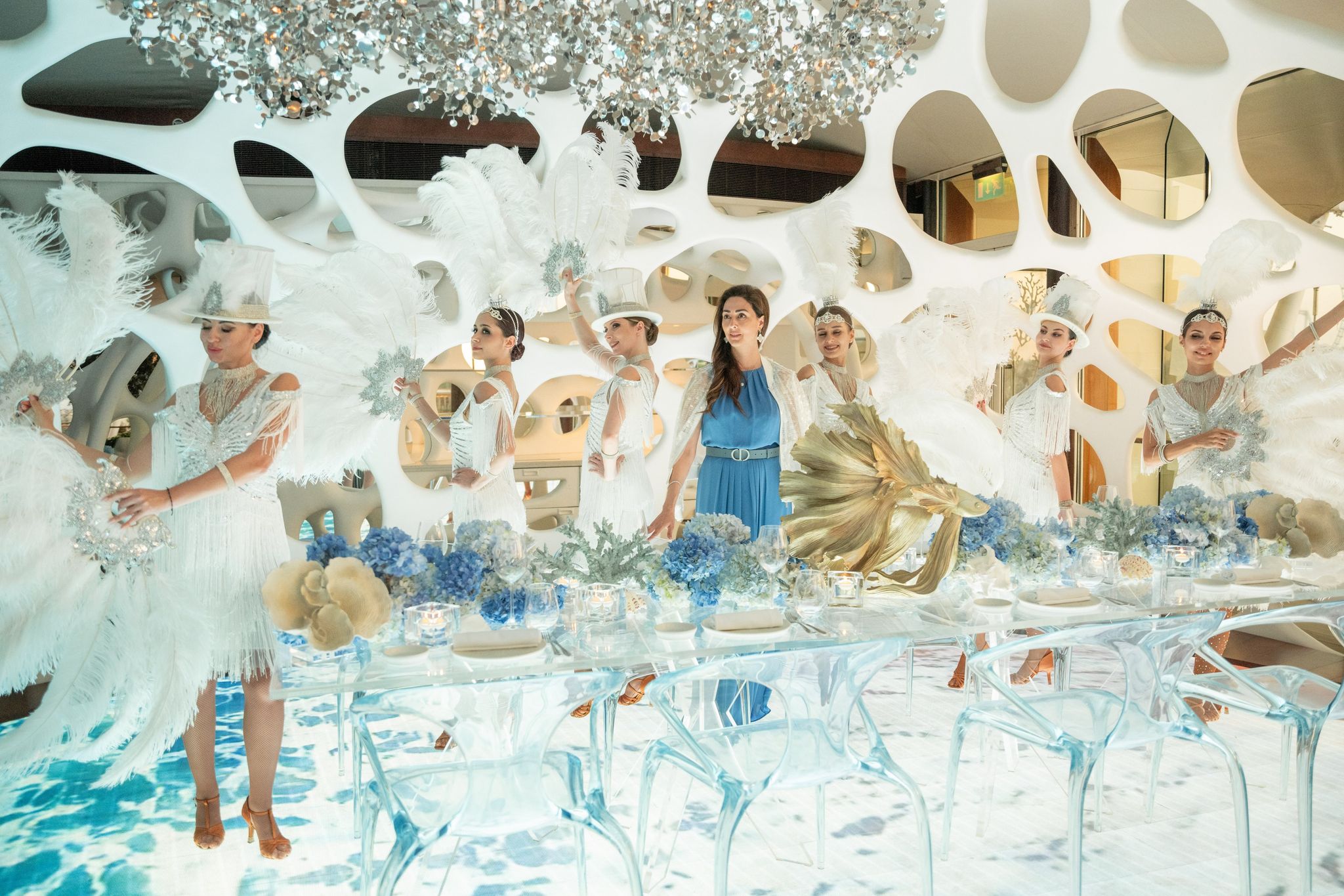 Feathered performers gathered around the long table beneath a glittering ceiling installation