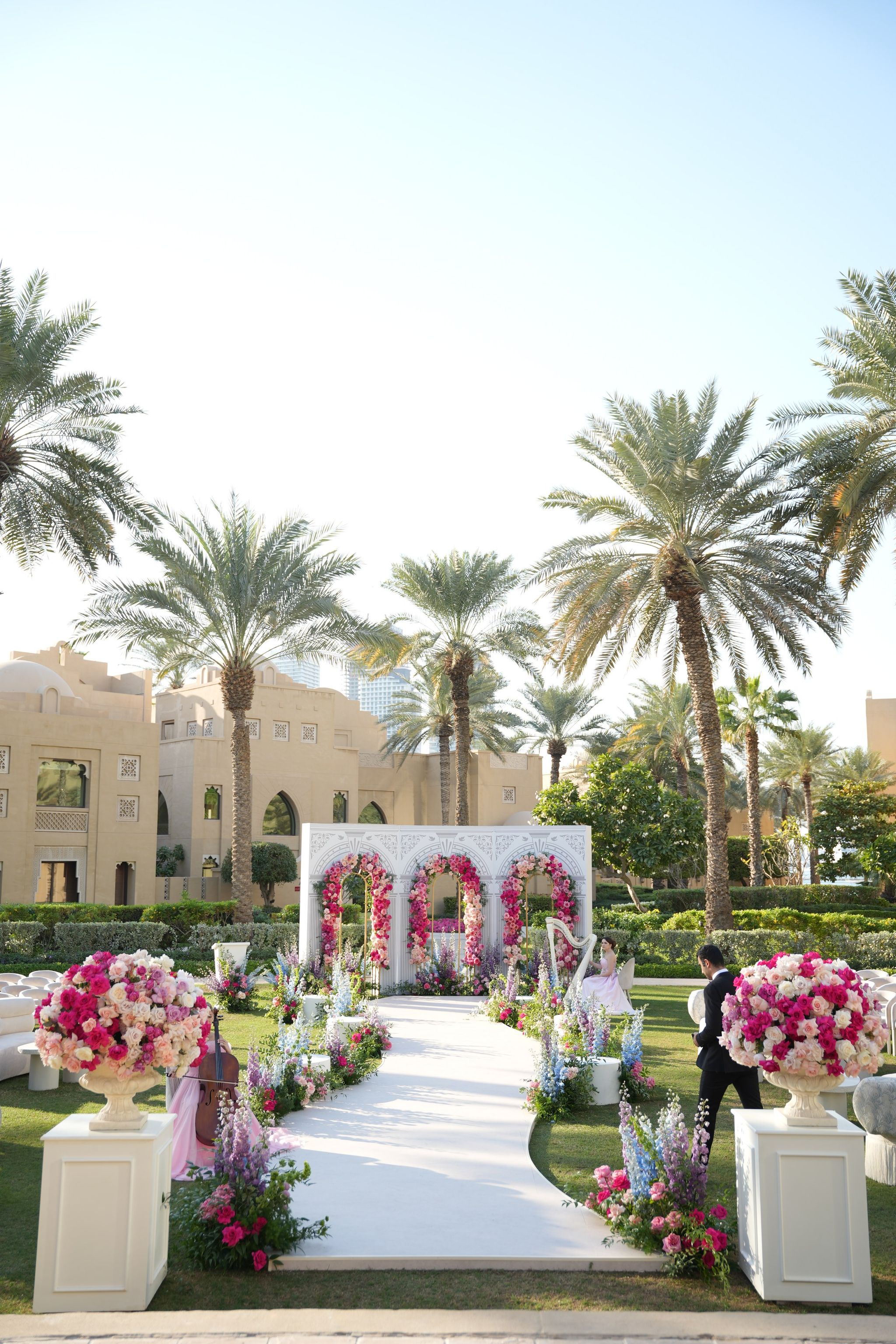 Ceremony aisle beneath tall date palms, flanked by urns of pink and white roses