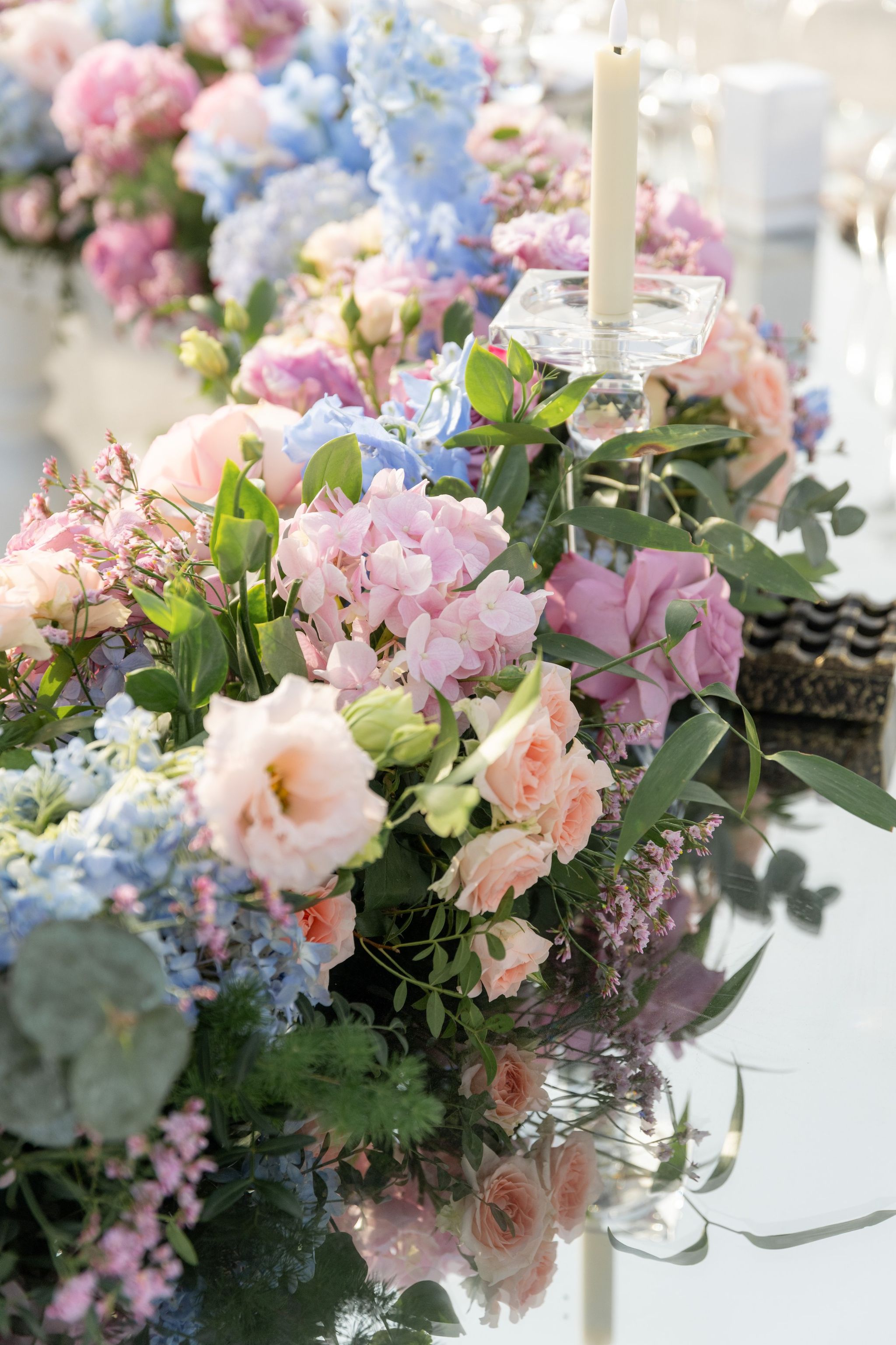 Low garland of blush roses, hydrangea and delphinium with a taper candle on a mirrored table