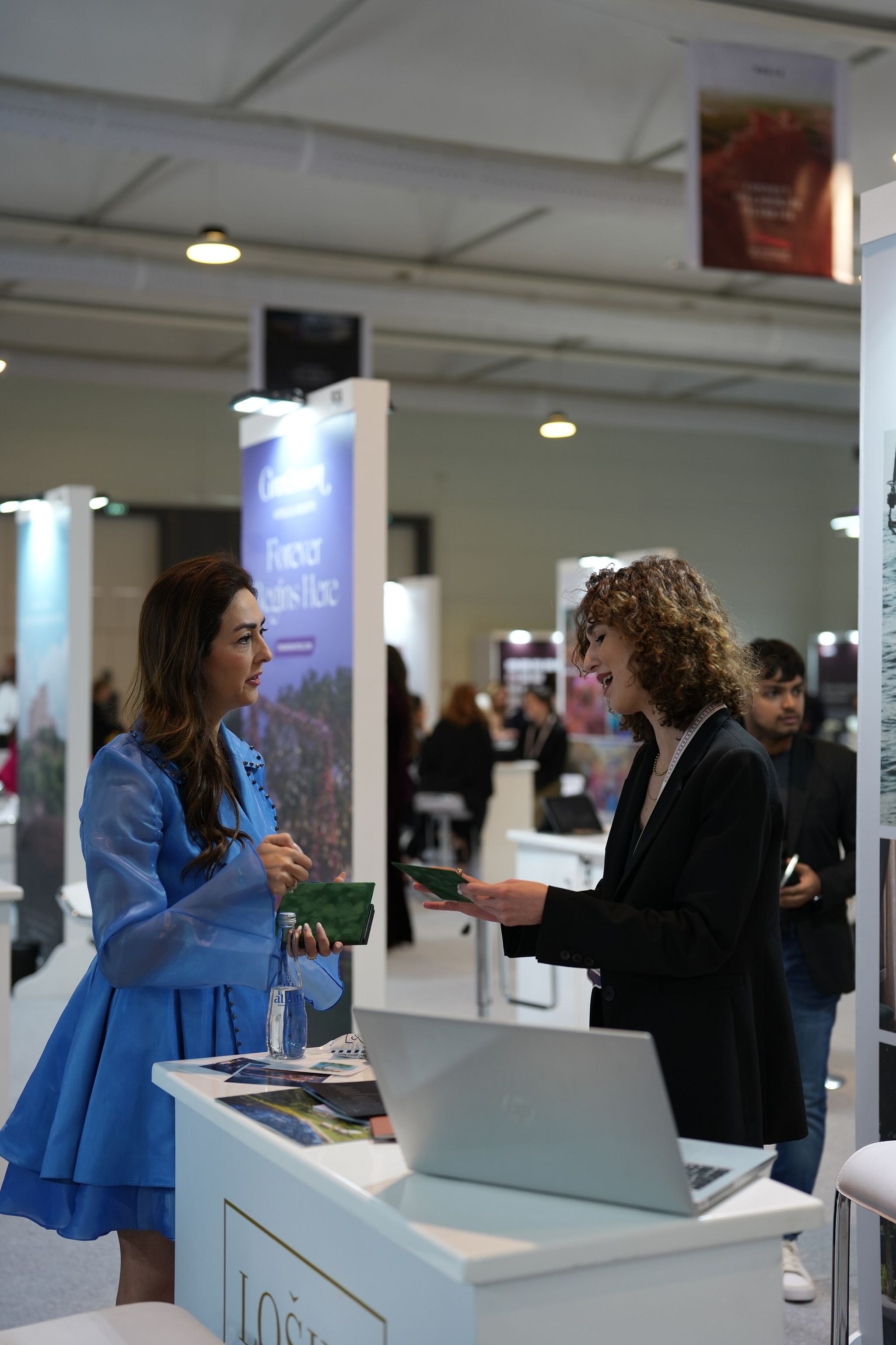 Exhibitors talking over a white counter with a laptop and brochures in a busy exhibition hall