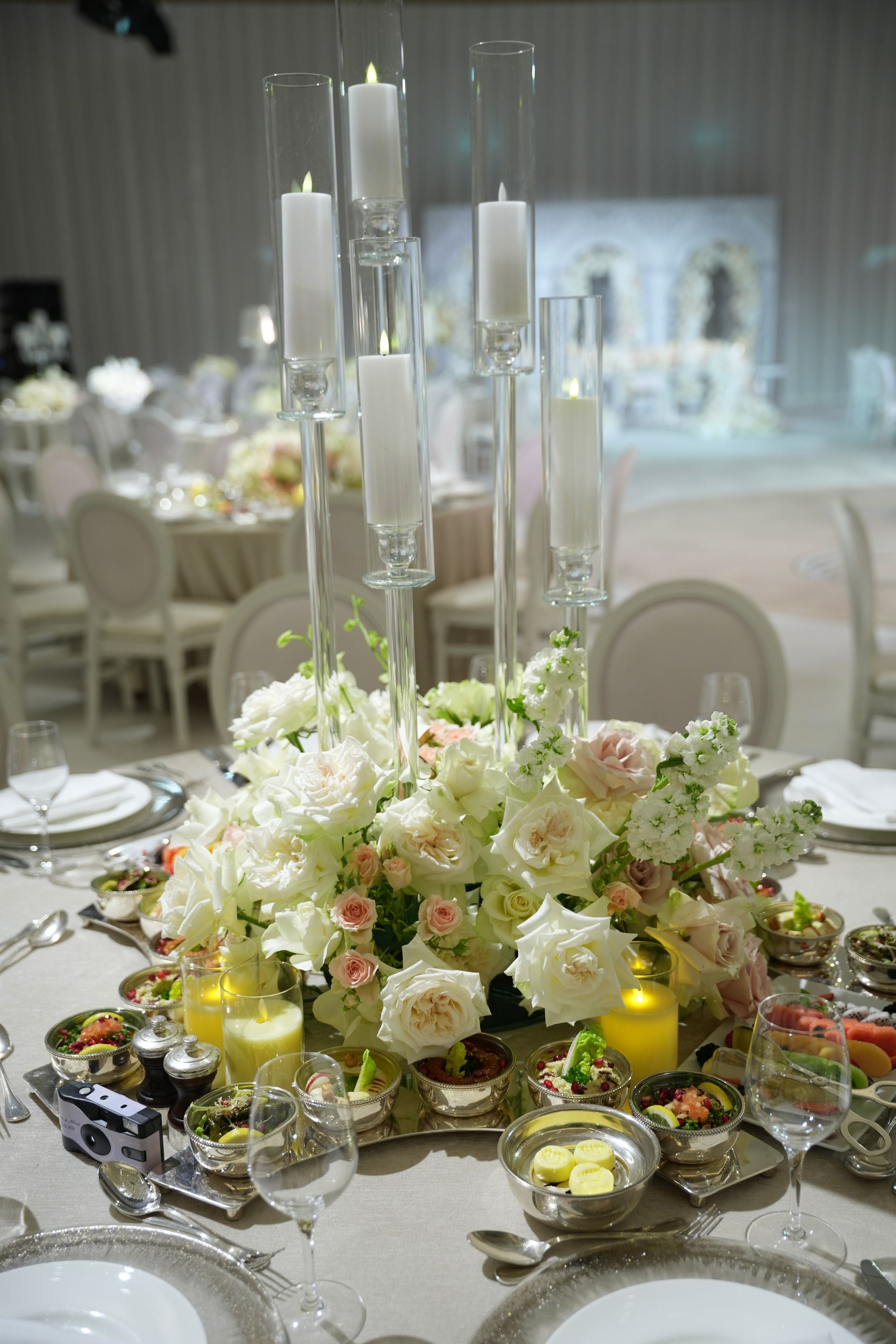 Tall glass candles over white roses, with small plates of starters served in silver bowls