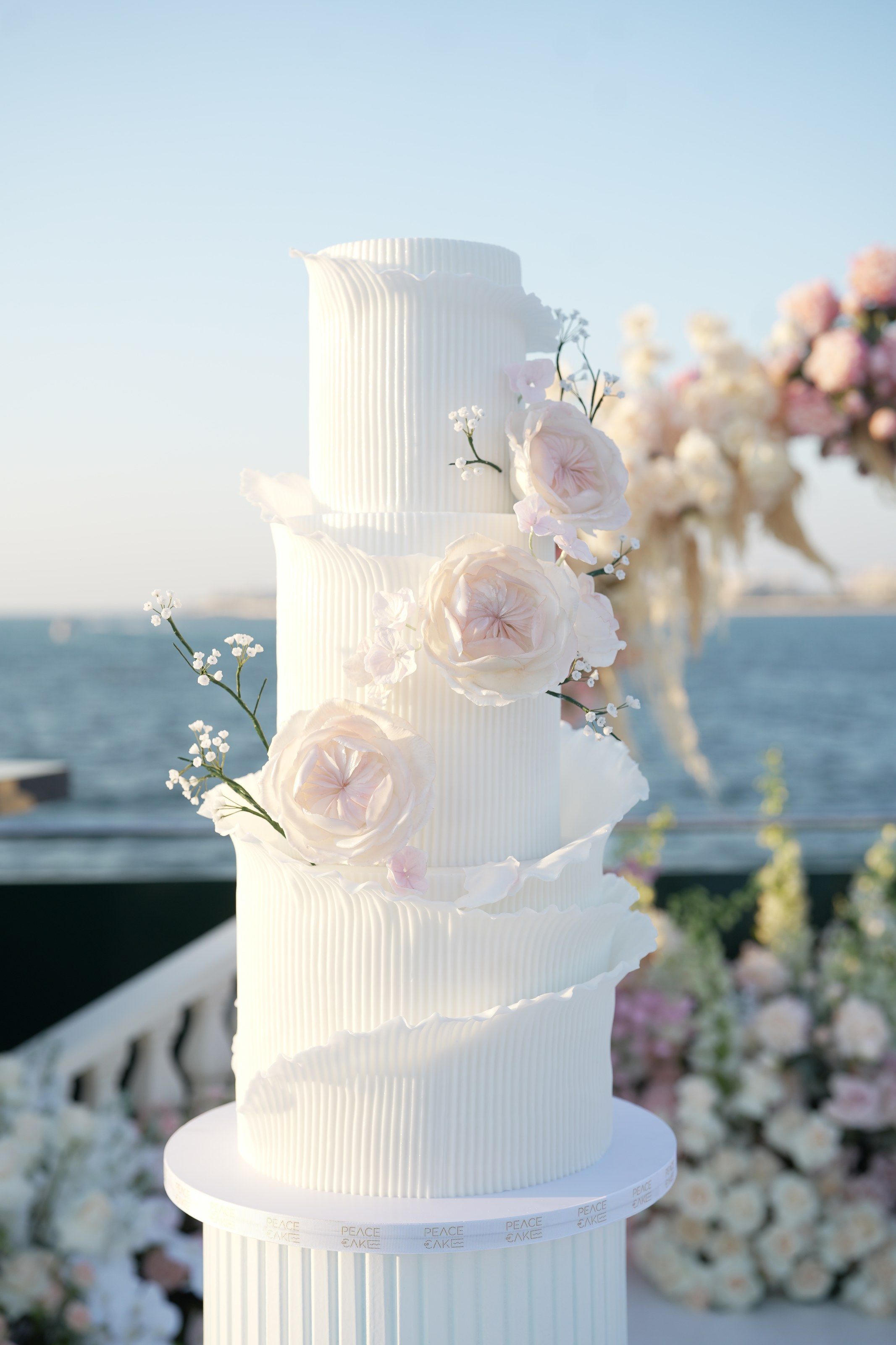 Tiered white cake with ribbed, ruffled layers and sugar roses against the sea at sunset