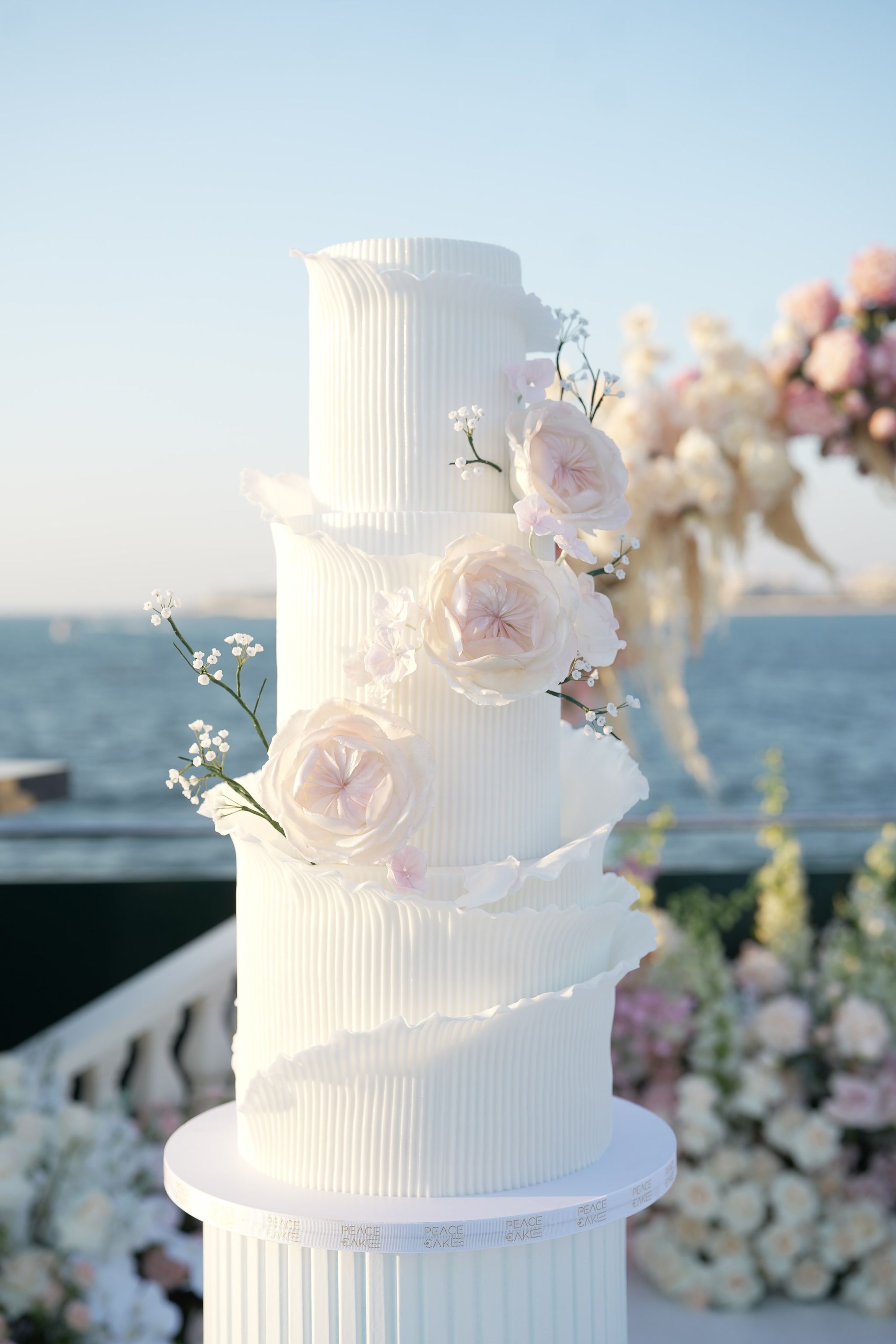 Tiered white cake with ribbed, ruffled layers and sugar roses against the sea at sunset