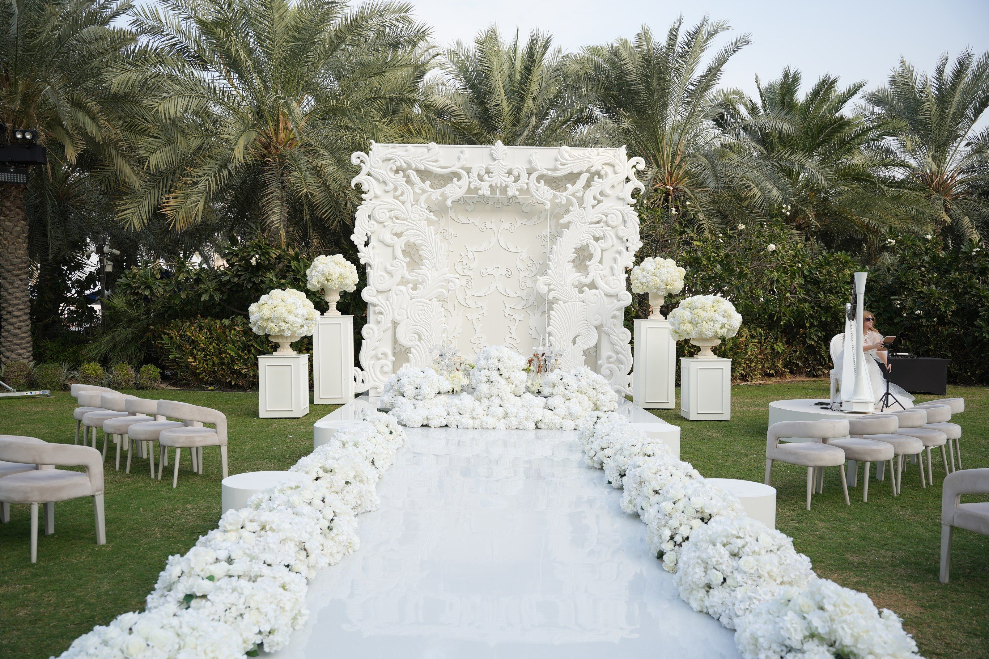 Carved white baroque ceremony backdrop and hydrangea-lined mirrored aisle on the palm-fringed lawn at Raffles The Palm Dubai