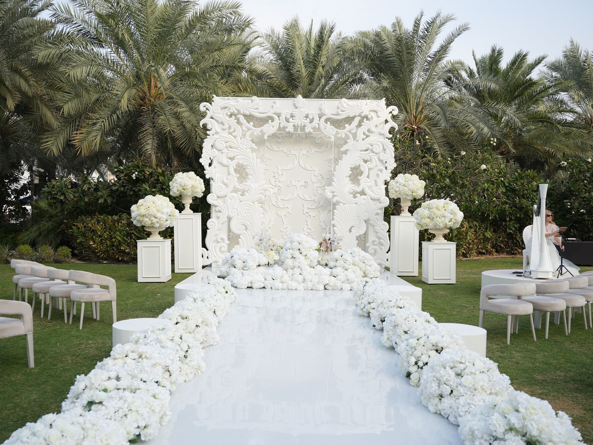 Carved white baroque ceremony backdrop and hydrangea-lined mirrored aisle on the palm-fringed lawn at Raffles The Palm Dubai