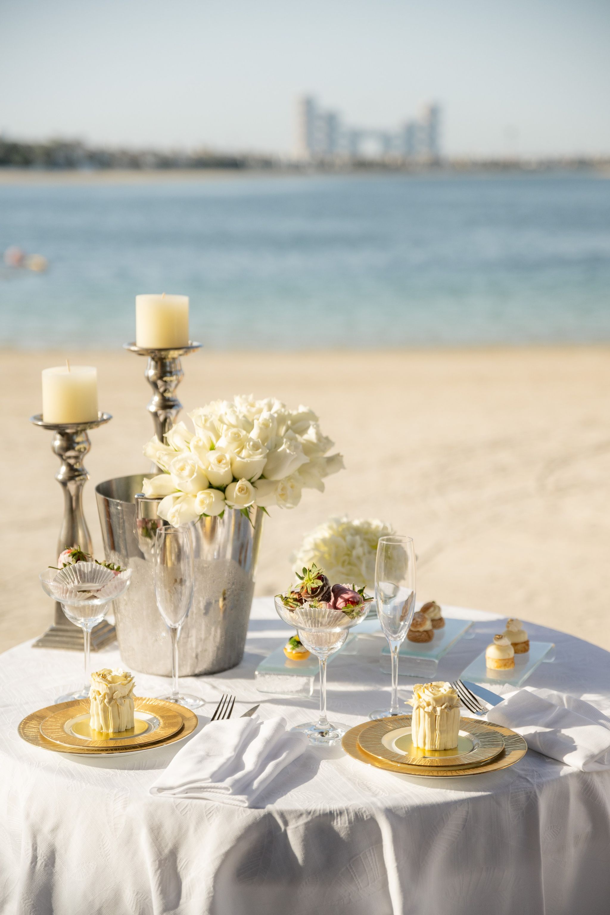 Beachside table for two with white roses, silver candlesticks and gold chargers, the sea beyond