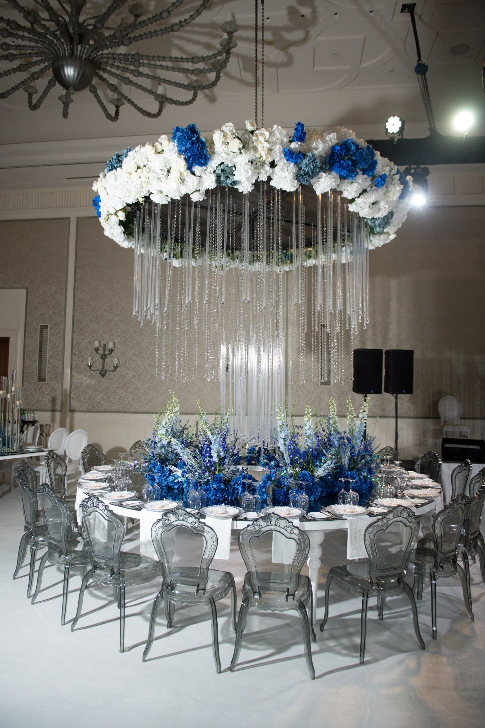 Round table under a ring of blue and white hydrangeas with cascading crystal strands and ghost chairs
