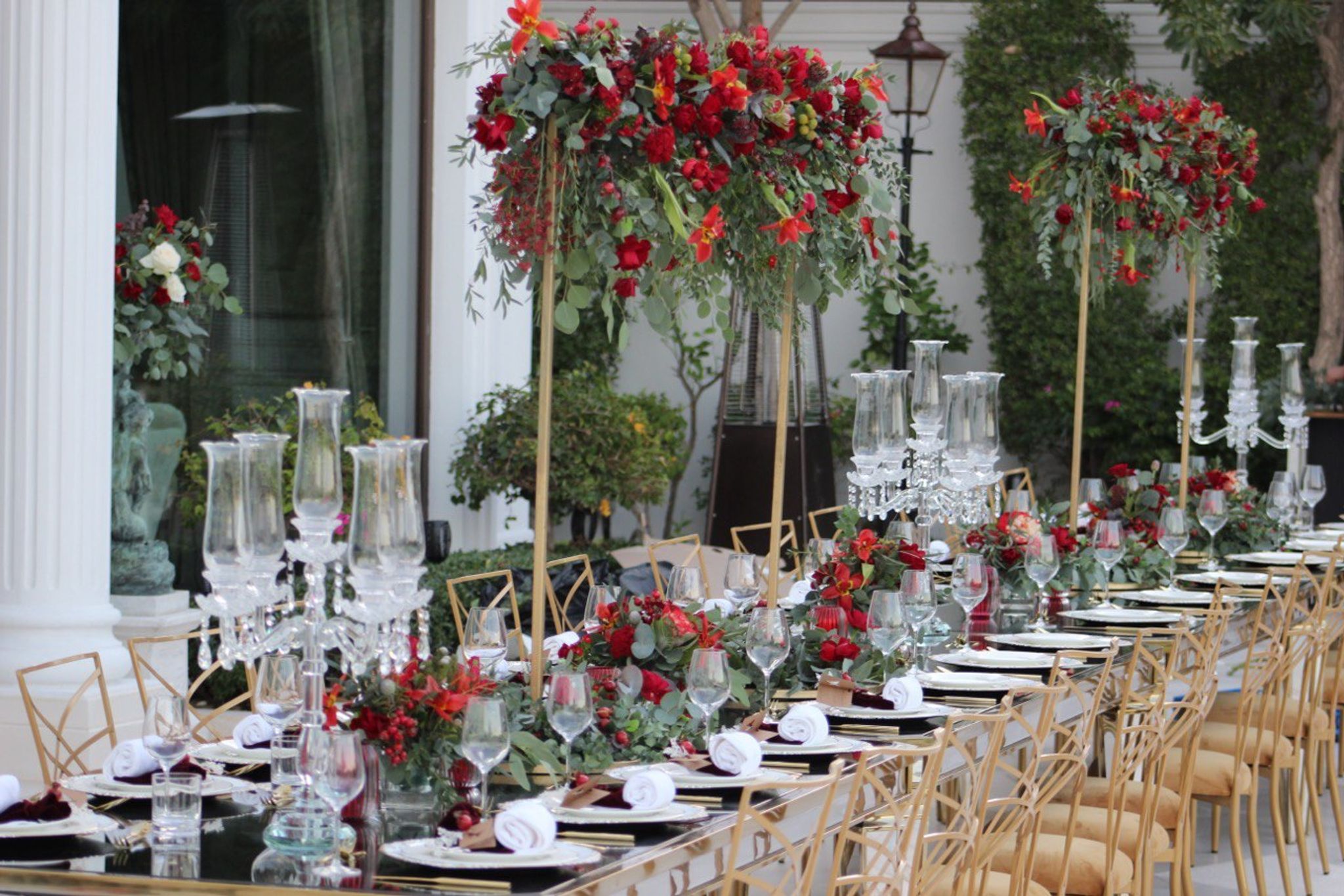 Red rose and lily arrangements on gold stands above a table of crystal candelabra and gold chairs
