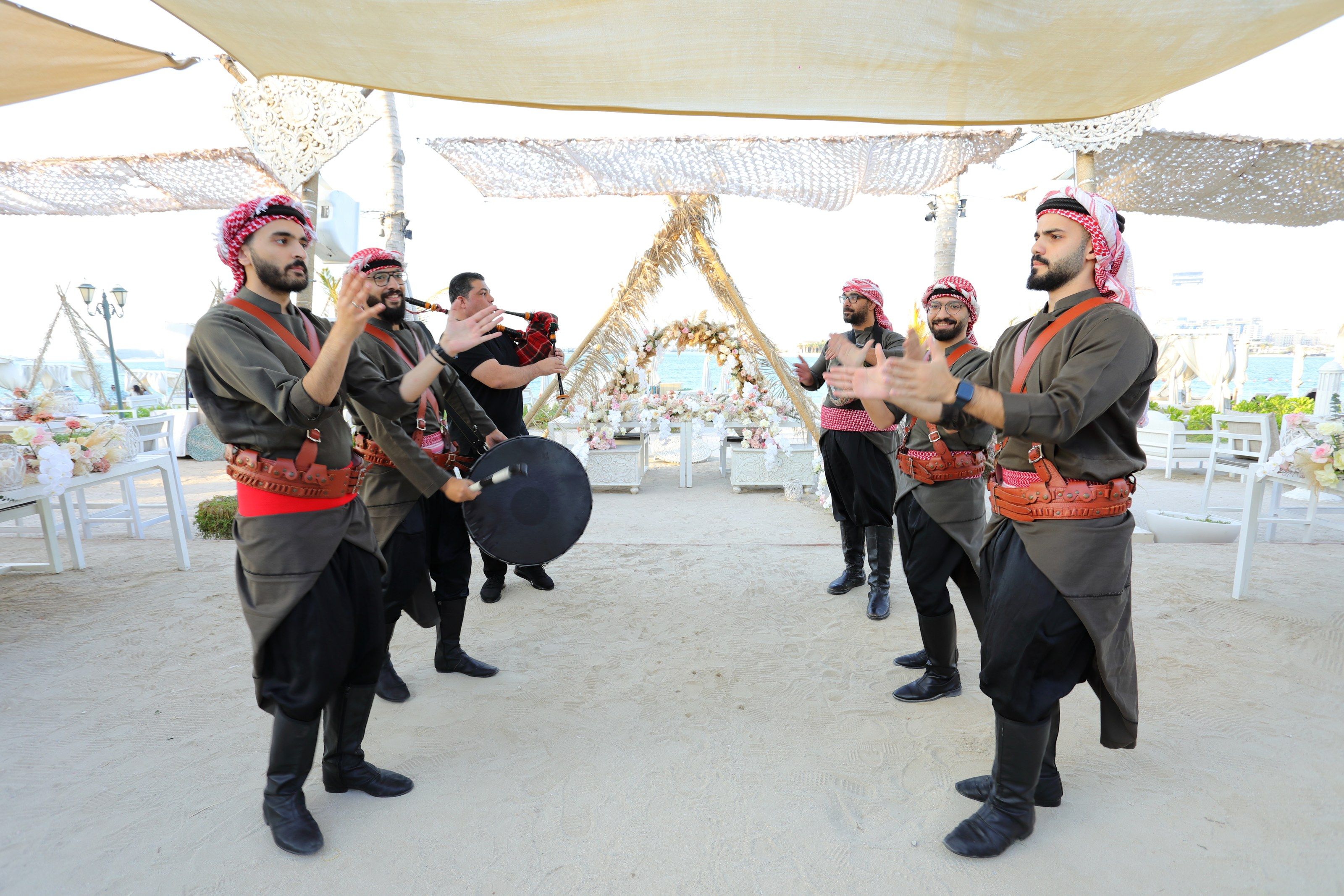 Traditional performers in keffiyehs with a drum and bagpipe beneath woven shade sails on the sand