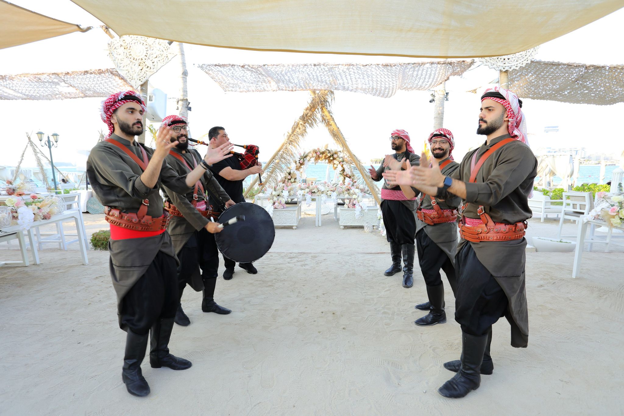 Traditional performers in keffiyehs with a drum and bagpipe beneath woven shade sails on the sand
