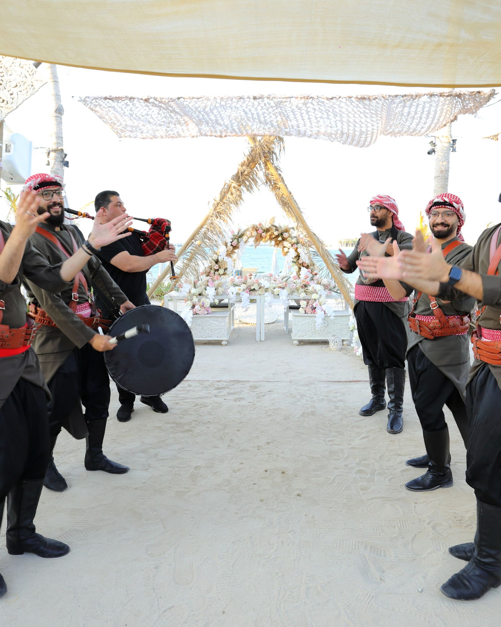 Traditional performers in keffiyehs with a drum and bagpipe beneath woven shade sails on the sand