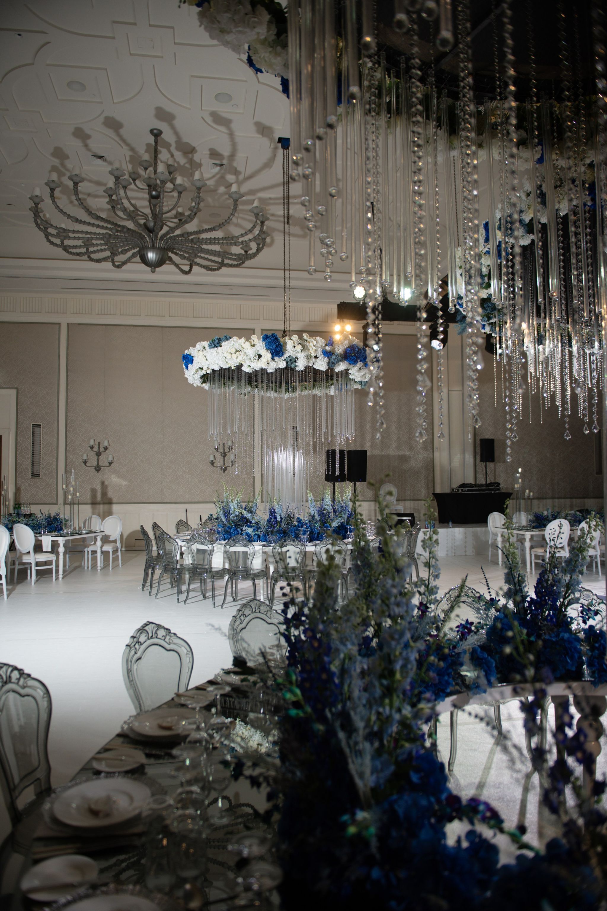 Crystal strands and blue delphiniums in the foreground, with floral rings and tables across the ballroom