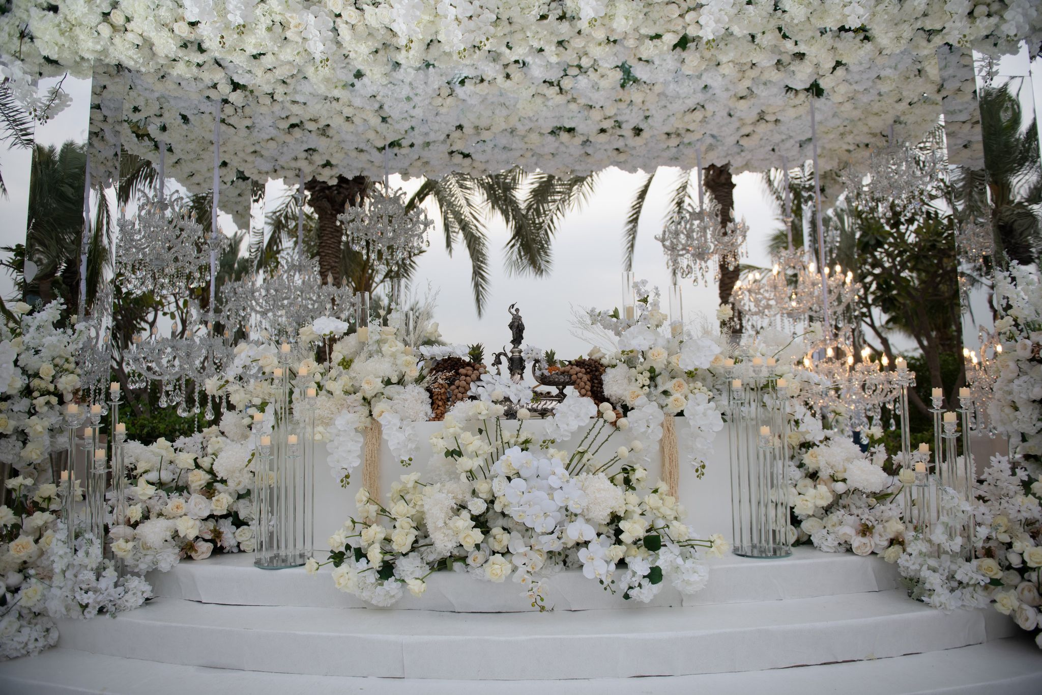 Stepped white stage under a canopy of white flowers and crystal chandeliers, sofreh at its centre