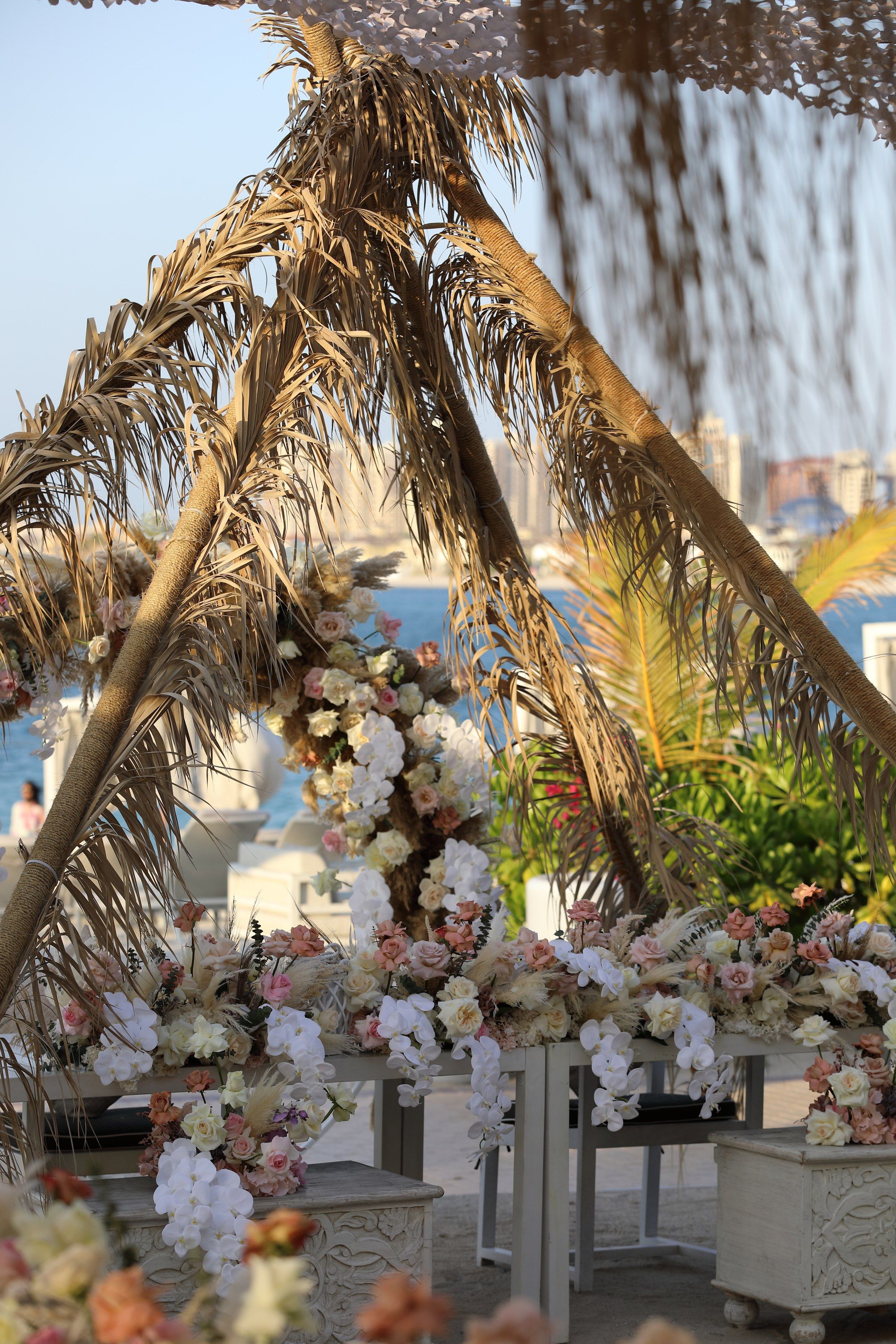 Dried palm-frond arch with cascading blush and ivory flowers, the sea and city towers beyond